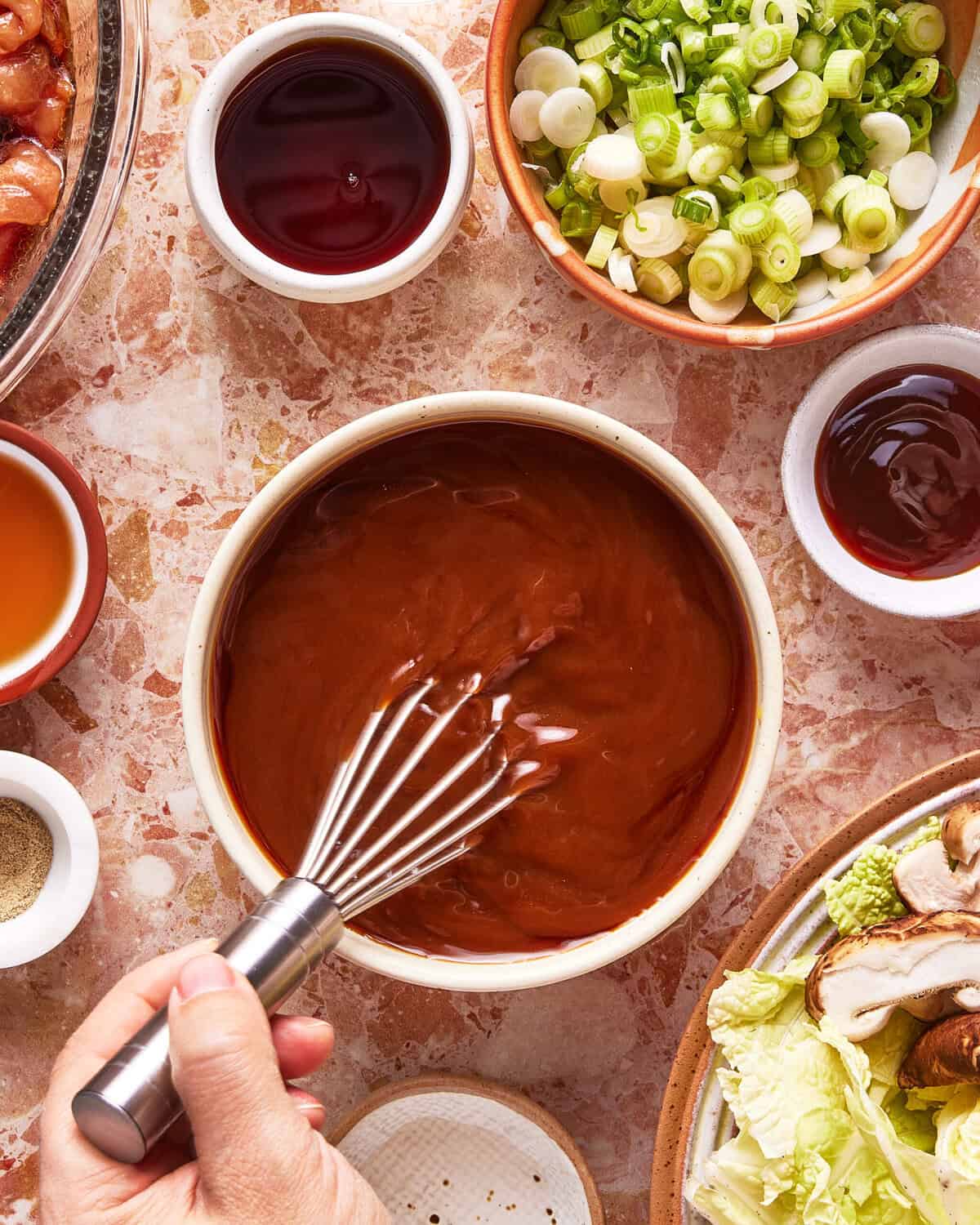 A hand using a whisk to stir a bowl of brown sauce, surrounded by small bowls containing sliced green onions, sauces, and vegetables on a marble surface.