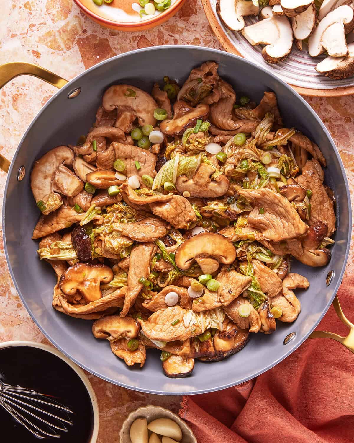 A skillet filled with stir-fried pork, mushrooms, and green onions, surrounded by bowls of sliced mushrooms, garlic cloves, and a small dish of dark sauce on a marble countertop.