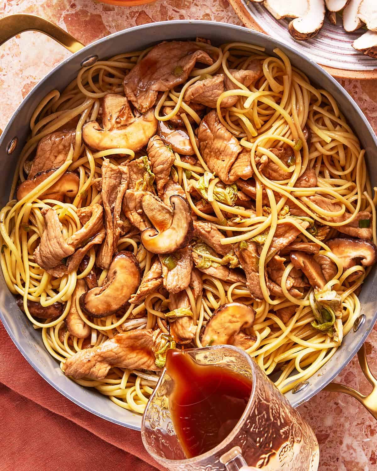 A skillet filled with cooked spaghetti, sliced mushrooms, strips of meat, and vegetables, with a glass measuring cup pouring a brown sauce over the pasta. A plate with extra mushroom slices is nearby.