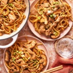 Two plates and a pot filled with stir-fried noodles, sliced beef, mushrooms, and green onions sit on a marble surface, with chopsticks and a glass of ice water nearby.
