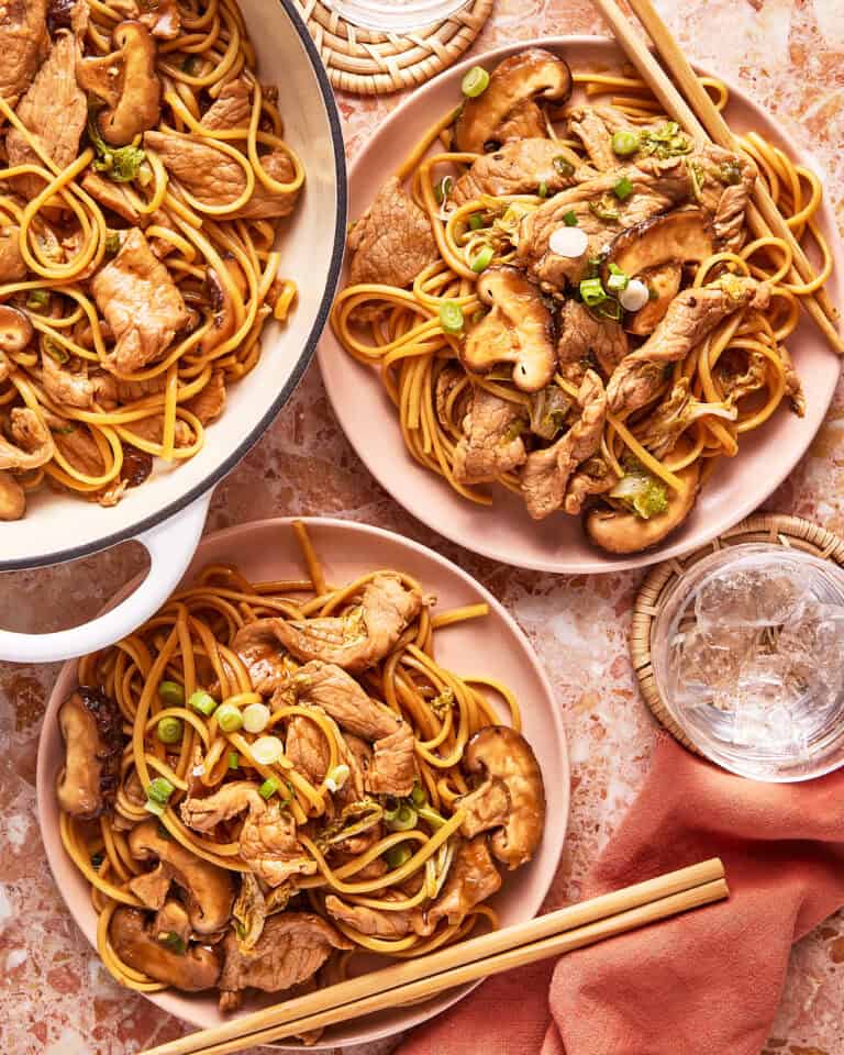Two plates and a pot filled with stir-fried noodles, sliced beef, mushrooms, and green onions sit on a marble surface, with chopsticks and a glass of ice water nearby.