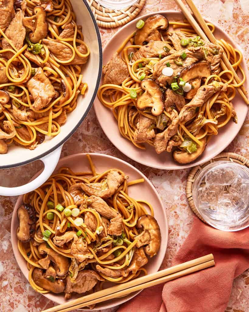 Two plates and a pot filled with stir-fried noodles, sliced beef, mushrooms, and green onions sit on a marble surface, with chopsticks and a glass of ice water nearby.