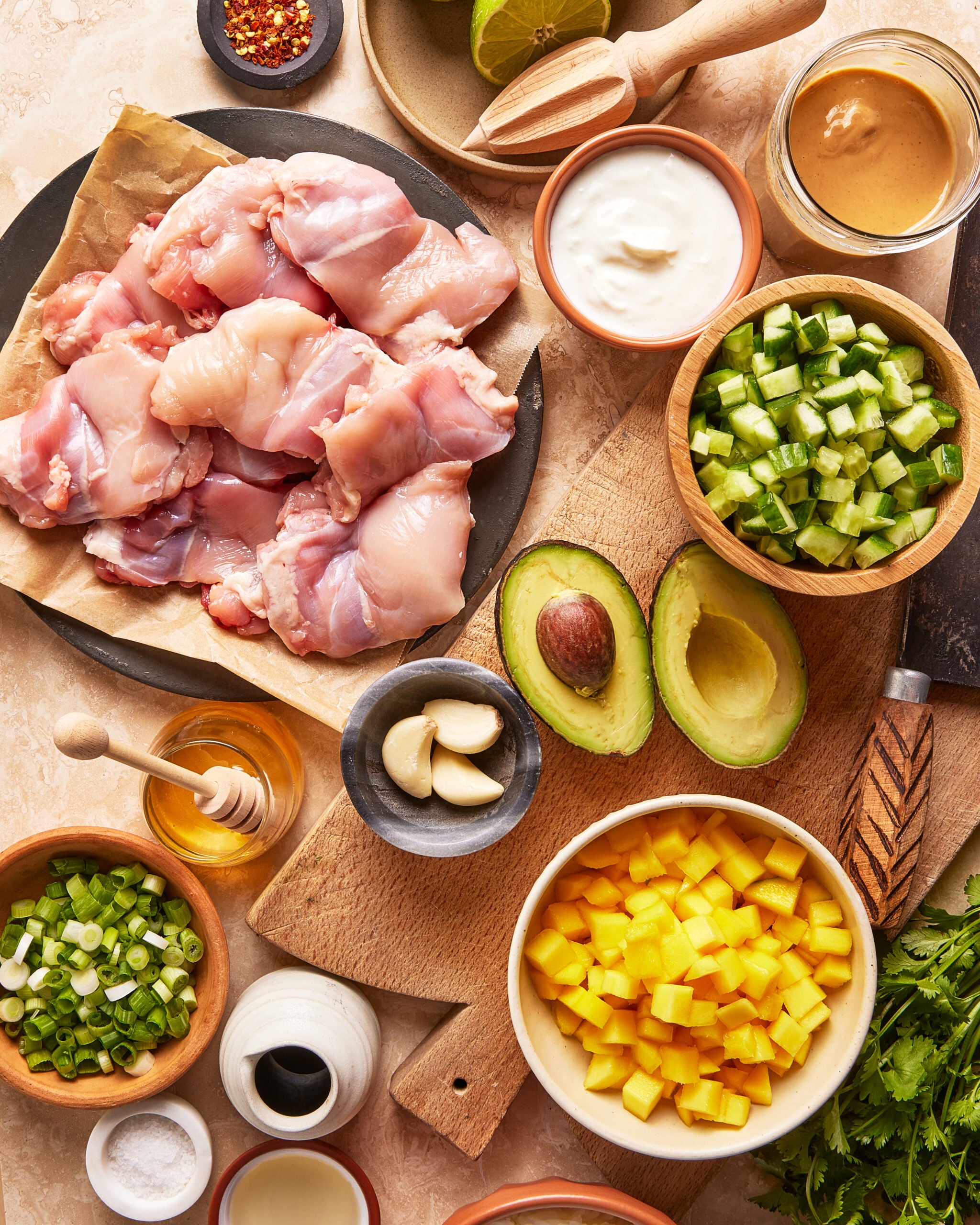 A top view of ingredients on a table, including raw chicken thighs, diced mango, chopped cucumber, sliced green onions, avocado halves, garlic cloves, sauces, honey, lime, spices, and fresh herbs.
