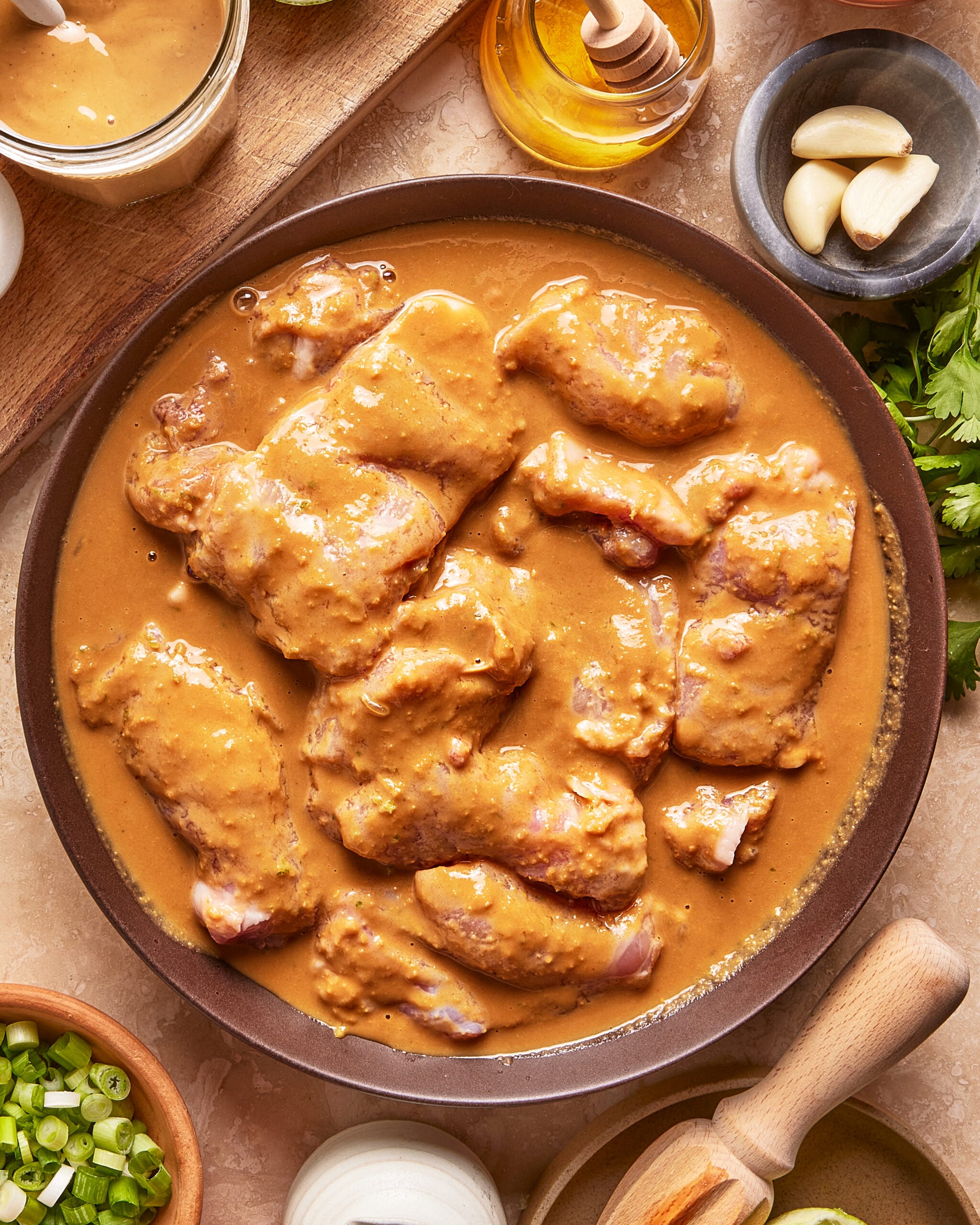 Raw chicken pieces coated in a thick, creamy brown marinade are arranged in a round dish. Surrounding the dish are fresh garlic cloves, green onions, parsley, a honey dipper, and various bowls with ingredients.