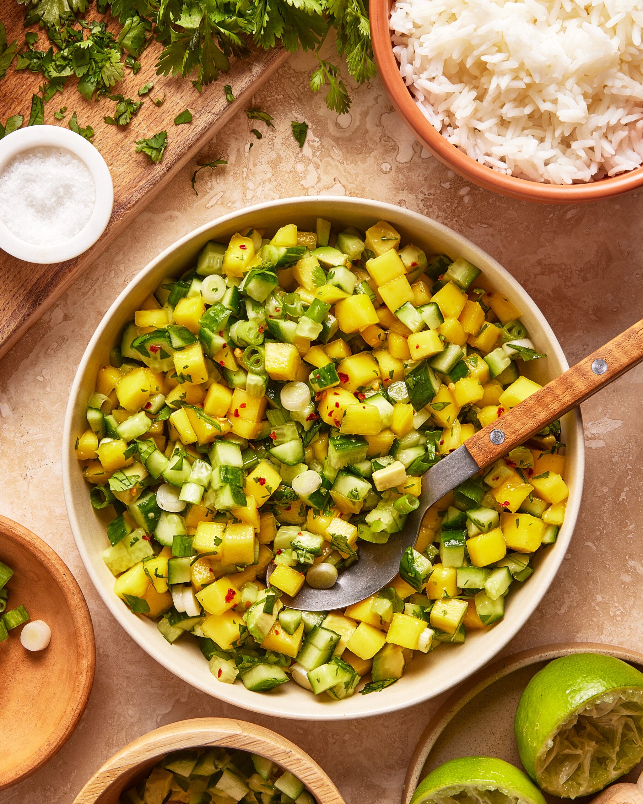 A bowl of mango cucumber salsa with chopped green onions, herbs, and spices sits on a table, surrounded by a wooden board with salt, chopped cilantro, a bowl of white rice, and halved limes.