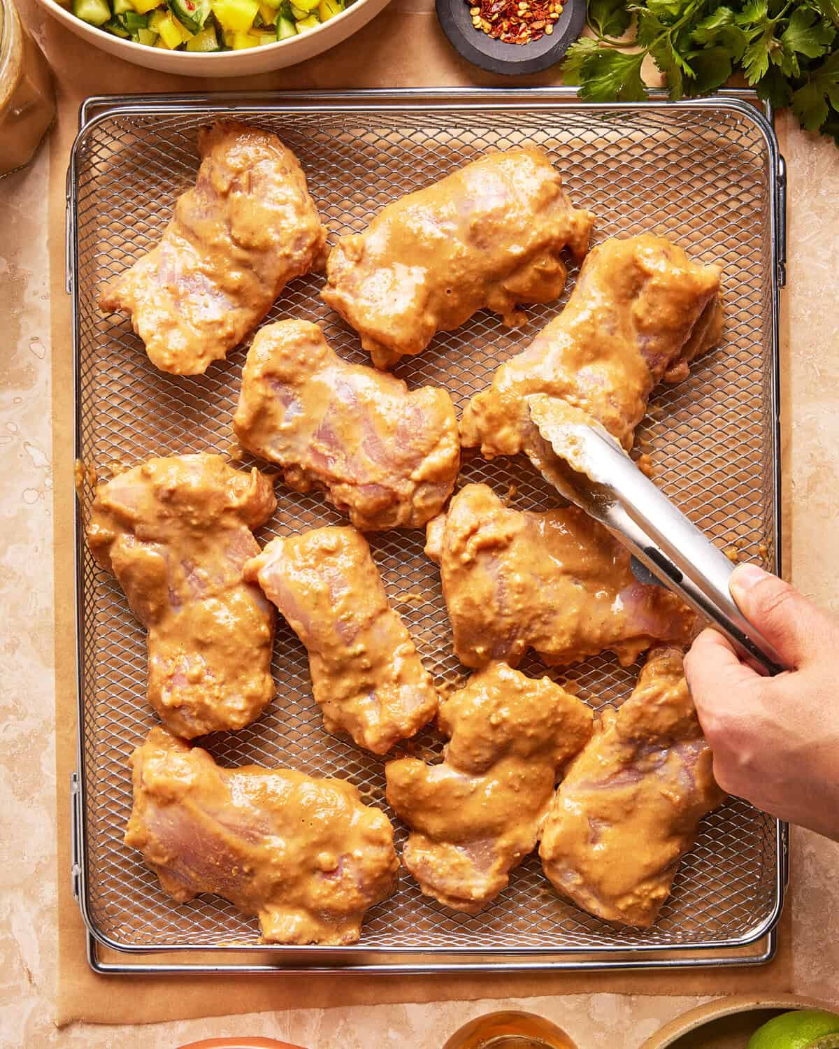 Raw, marinated chicken thighs arranged on an air fryer rack. A hand with metal tongs is placing or adjusting one piece. Fresh herbs and a chopped vegetable mixture are partially visible nearby.