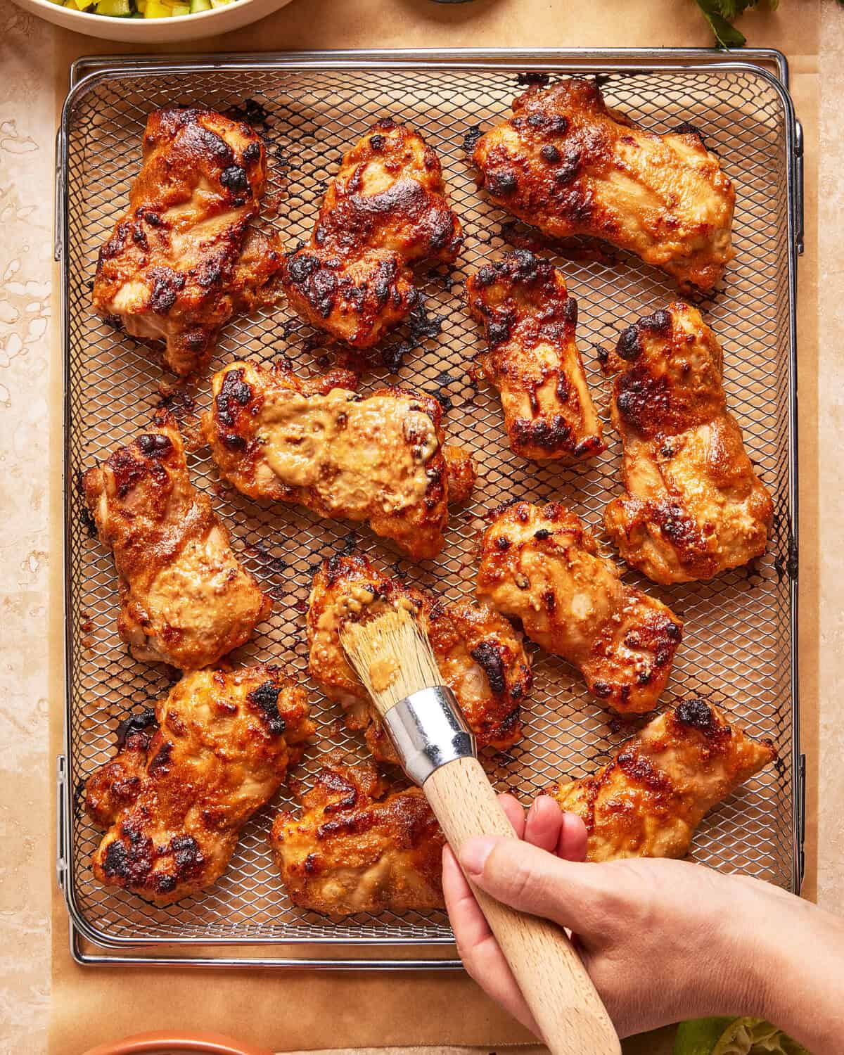 A hand uses a brush to apply sauce to grilled chicken pieces on a wire rack, with a few char marks visible on the meat.