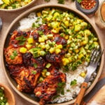 A bowl of grilled chicken thighs served over white rice, topped with a fresh mango-cucumber salsa. A fork rests on the plate, with cilantro, chopped green onions, and red pepper flakes nearby.