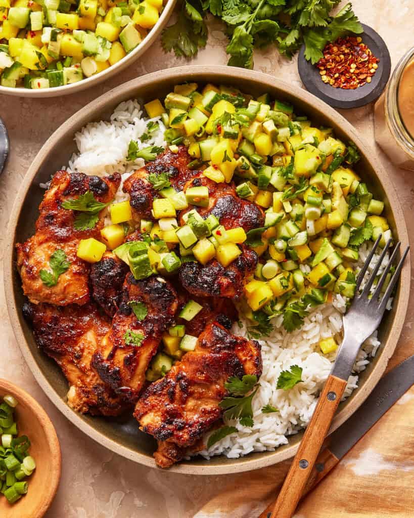 A bowl of grilled chicken thighs served over white rice, topped with a fresh mango-cucumber salsa. A fork rests on the plate, with cilantro, chopped green onions, and red pepper flakes nearby.