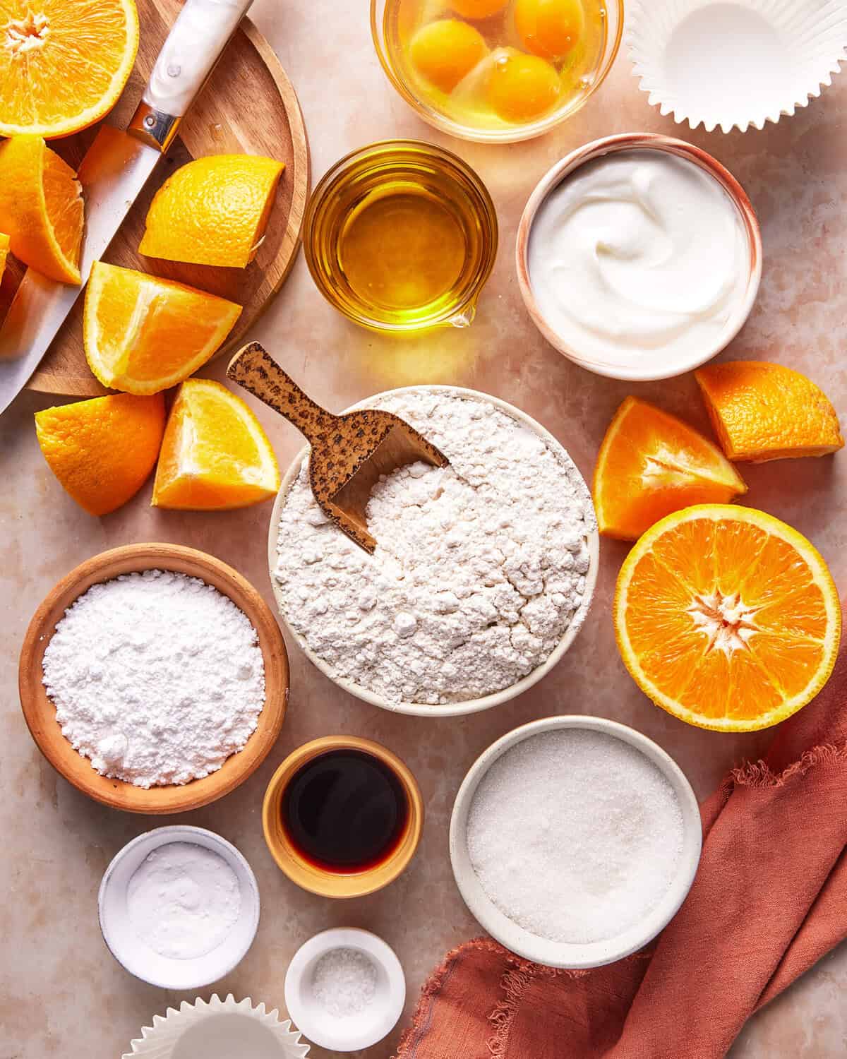 A flat lay of baking ingredients including flour, sugar, powdered sugar, eggs, yogurt, vanilla extract, orange slices, olive oil, and baking powder on a light surface with a wooden spoon and napkin.