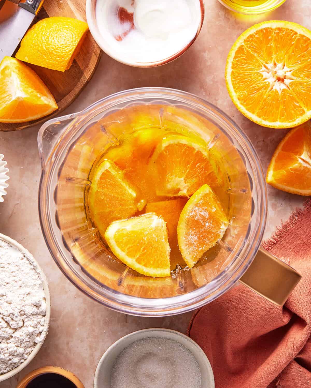 A glass bowl with orange wedges submerged in liquid, surrounded by halved oranges, flour, sugar, a small bowl of yogurt, and an orange napkin on a marble countertop.