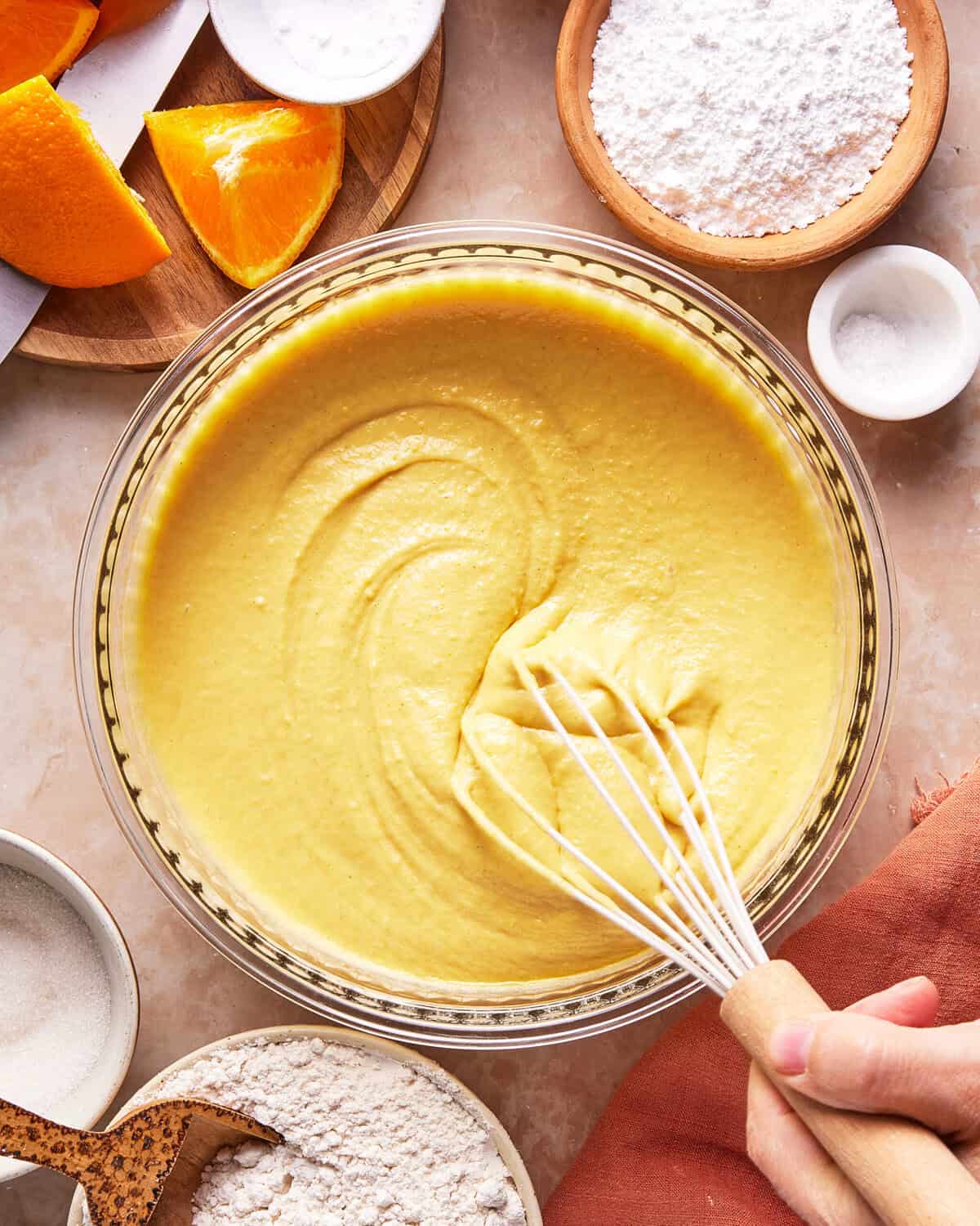 A hand whisking smooth yellow cake batter in a glass bowl, surrounded by orange slices, powdered sugar, flour, and sugar on a countertop.