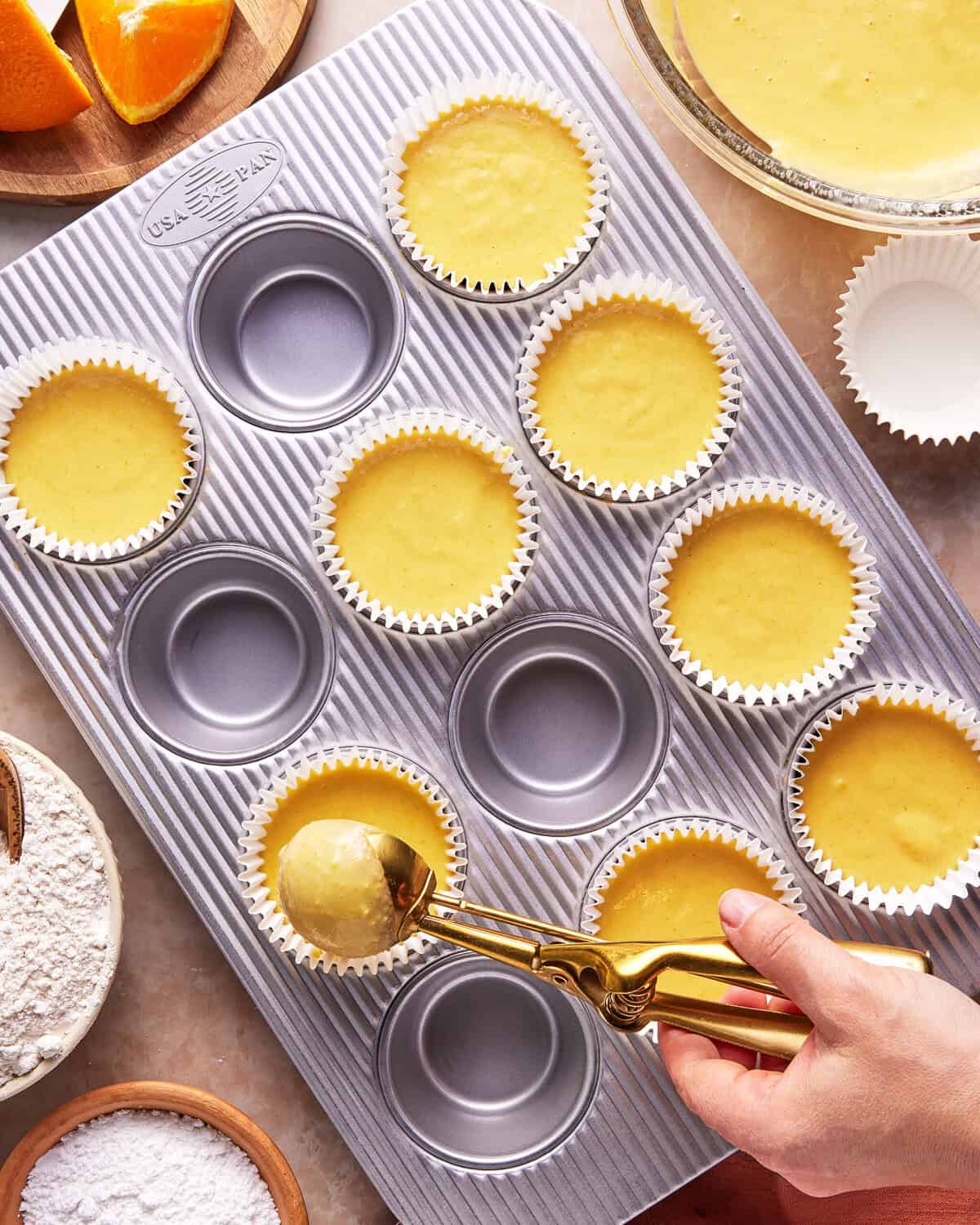 A hand uses a scoop to fill cupcake liners in a muffin tray with yellow batter. Nearby are bowls of flour, batter, and orange slices on a wooden board.