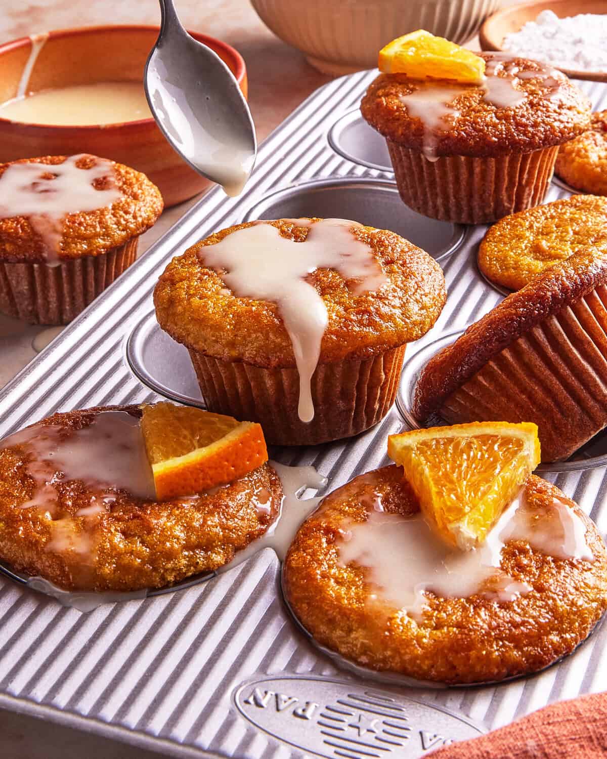 A muffin tin holds moist muffins drizzled with glaze, some topped with orange slices. A spoon pours glaze onto a muffin, while other baking items are visible in the background.