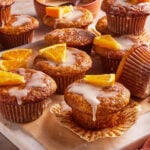 A batch of glazed muffins topped with fresh orange slices is arranged on a marble slab, with some muffins unwrapped and a bowl of glaze in the background.