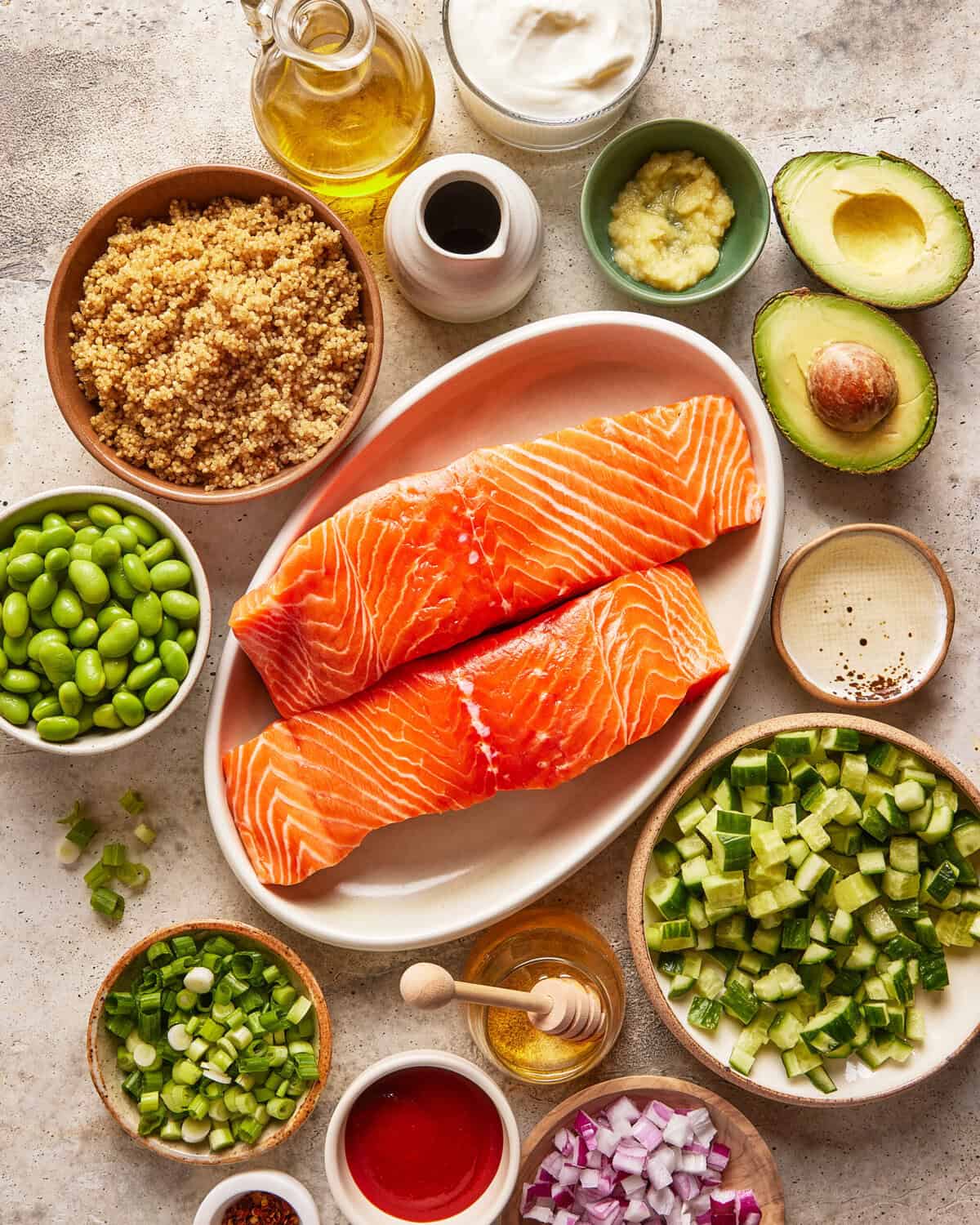 Two raw salmon fillets on a plate surrounded by bowls of ingredients including quinoa, edamame, diced cucumber, red onion, avocado, green onions, garlic, oil, vinegar, and sauces on a light surface.