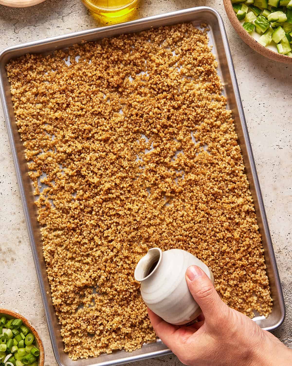 A hand pours liquid from a small white pitcher over a baking sheet covered with cooked quinoa. Surrounding the tray are bowls containing chopped green vegetables and a jar of yellow oil.