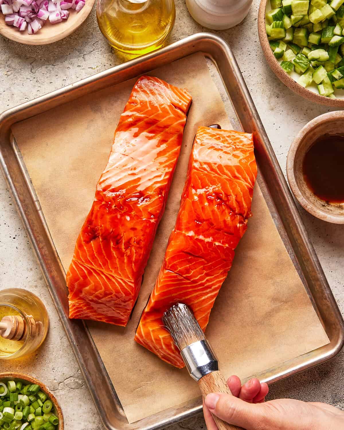 Two raw salmon fillets on a parchment-lined baking tray being brushed with marinade; surrounded by small bowls of chopped onions, cucumbers, oil, and sauce.