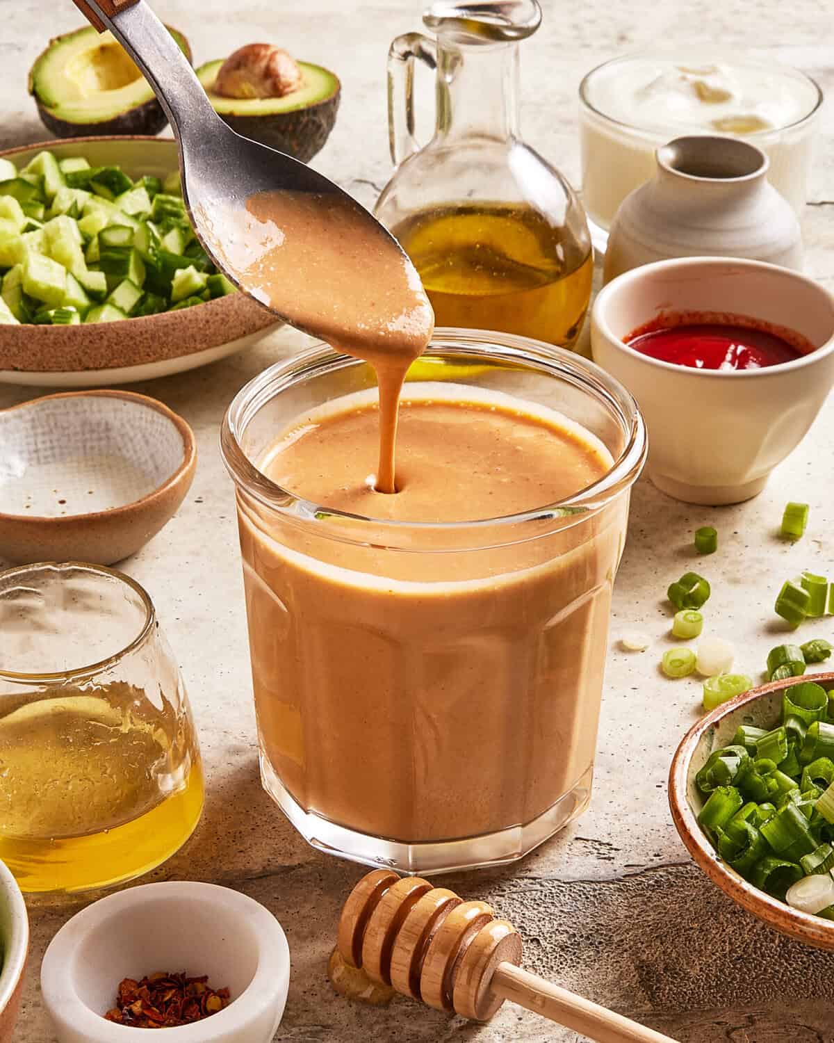 A spoon drizzles creamy, brown sauce into a glass surrounded by bowls of chopped cucumber, green onions, oil, red sauce, honey, and other fresh ingredients on a light countertop.