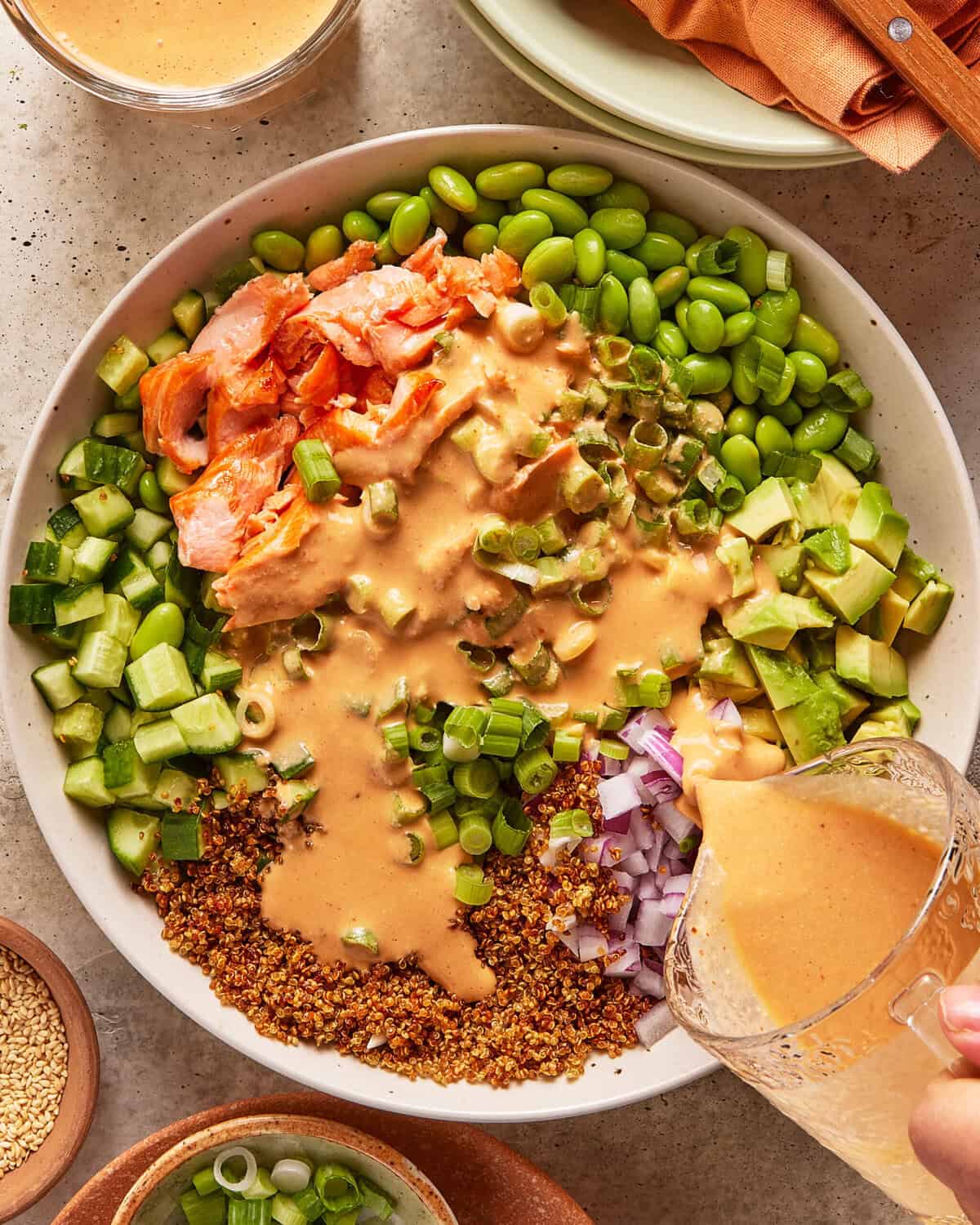 A bowl filled with diced cucumber, edamame, avocado, flaked salmon, red onion, green onion, crunchy topping, and creamy dressing being poured over the ingredients. Nearby are bowls with sesame seeds and extra dressing.