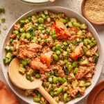 A bowl of salmon rice salad with chunks of salmon, edamame, diced cucumber, scallions, and sprinkled sesame seeds. A wooden spoon rests in the bowl, and a small dish of sesame seeds is nearby.