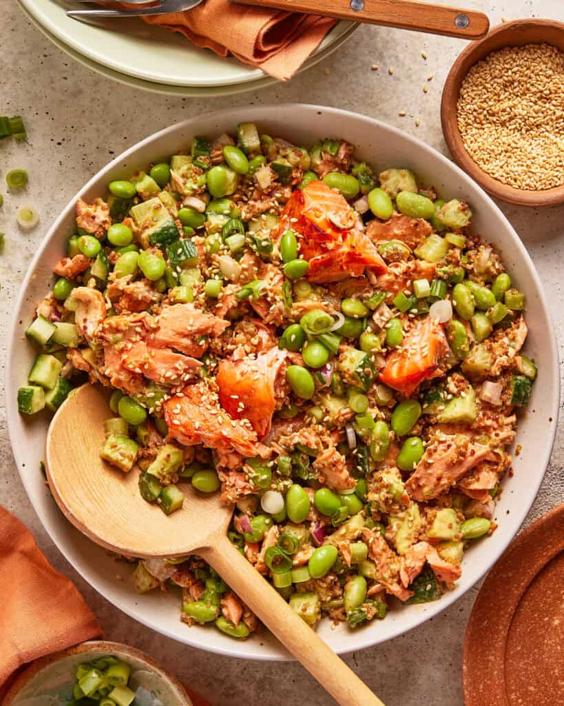 A bowl of salmon rice salad with chunks of salmon, edamame, diced cucumber, scallions, and sprinkled sesame seeds. A wooden spoon rests in the bowl, and a small dish of sesame seeds is nearby.