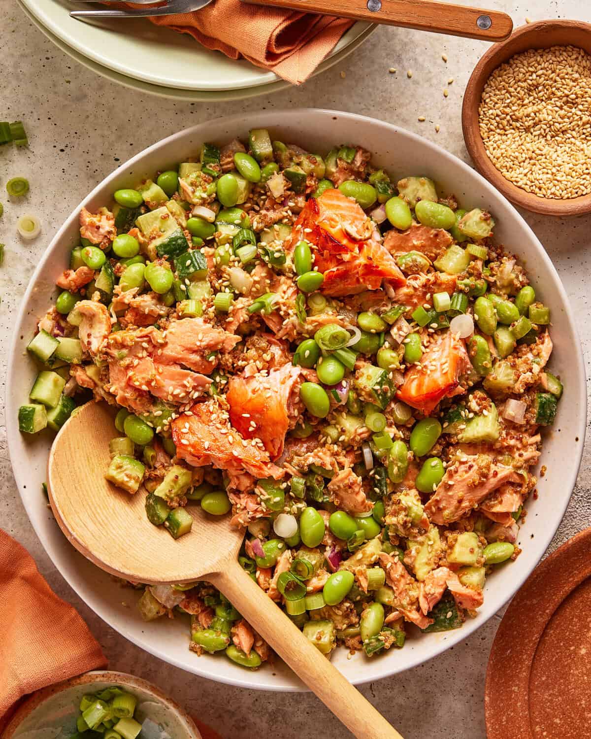 A bowl of salmon rice salad with chunks of salmon, edamame, diced cucumber, scallions, and sprinkled sesame seeds. A wooden spoon rests in the bowl, and a small dish of sesame seeds is nearby.