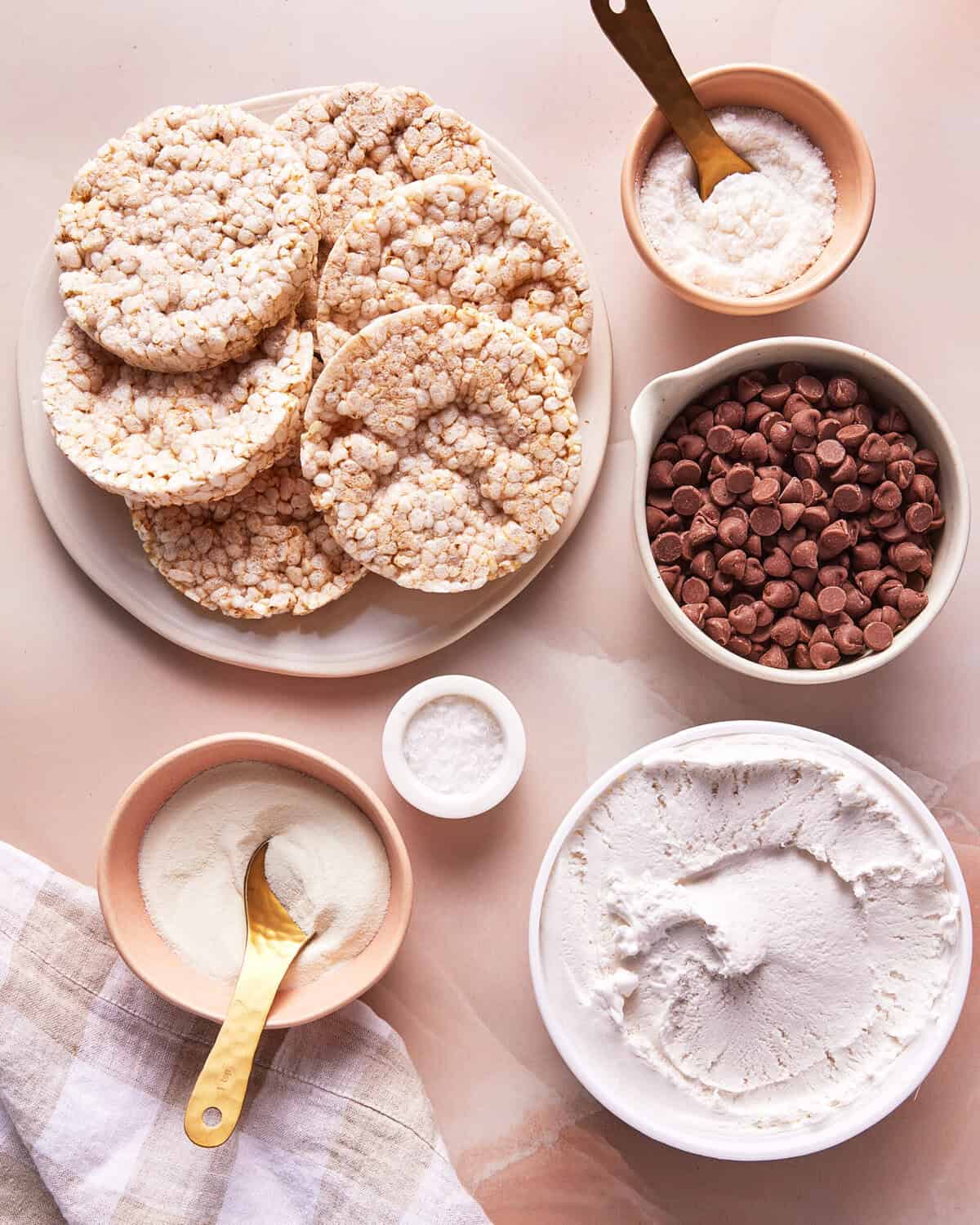 A flat lay of round rice cakes on a plate, chocolate chips in a bowl, a bowl of whipped cream, a bowl of powdered sugar with a gold spoon, a small bowl of salt, and a striped cloth on a light surface.