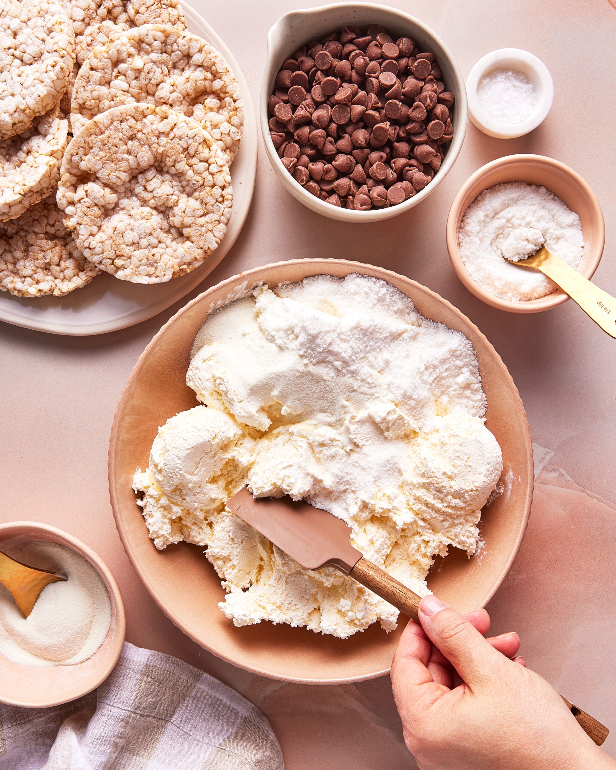 A hand spreads whipped cream in a bowl with a spatula. Surrounding bowls contain chocolate chips, sugar, and salt, with round rice cakes arranged on a plate nearby. All items are on a light-colored surface.