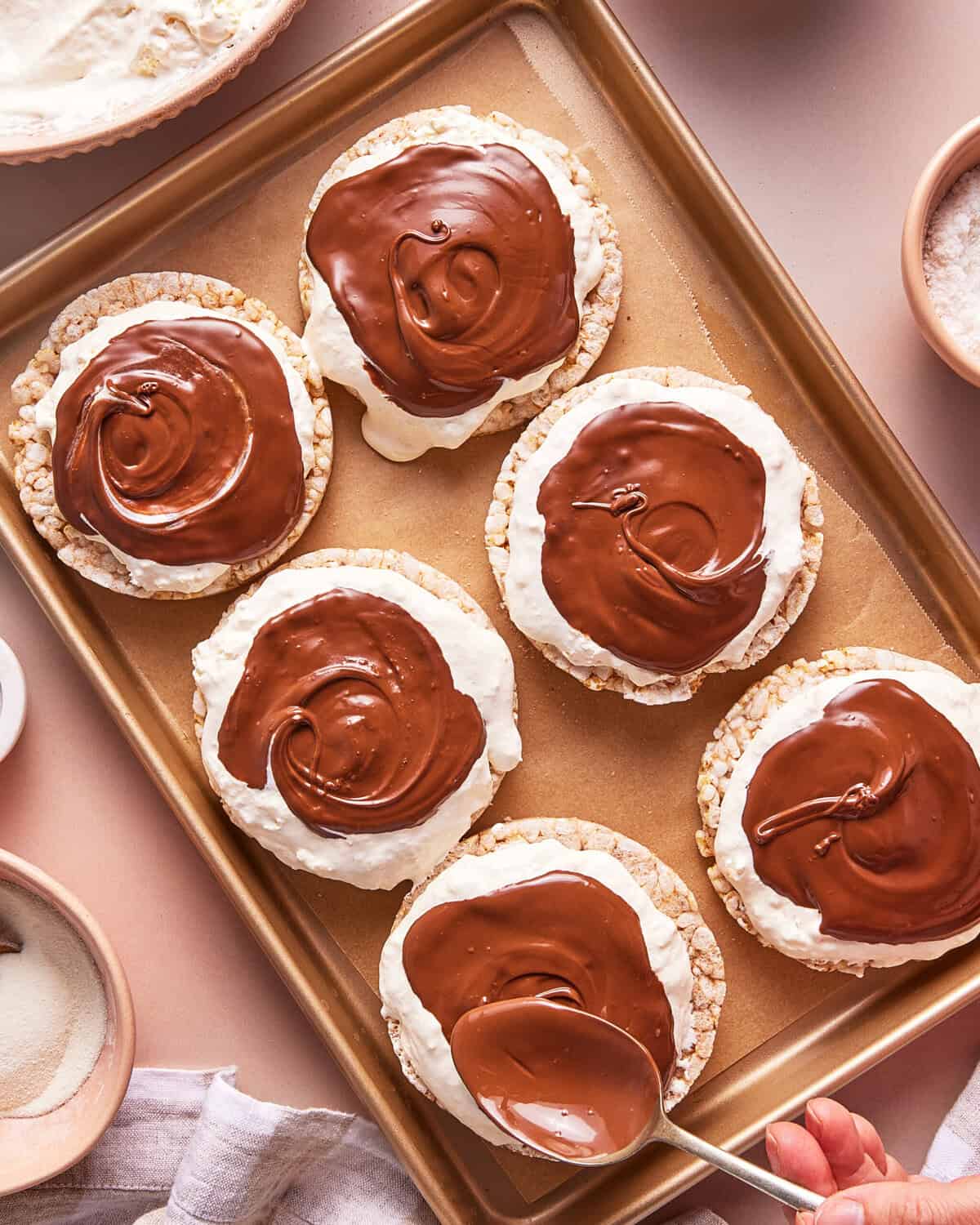 A hand spreads melted chocolate over rice cakes topped with a layer of creamy spread, arranged on a gold baking sheet with bowls of ingredients nearby.