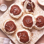 Round rice cakes topped with fluffy white cream and swirled melted chocolate, sprinkled with sea salt flakes, are arranged on a speckled cutting board. A napkin and bowls of ingredients are nearby.