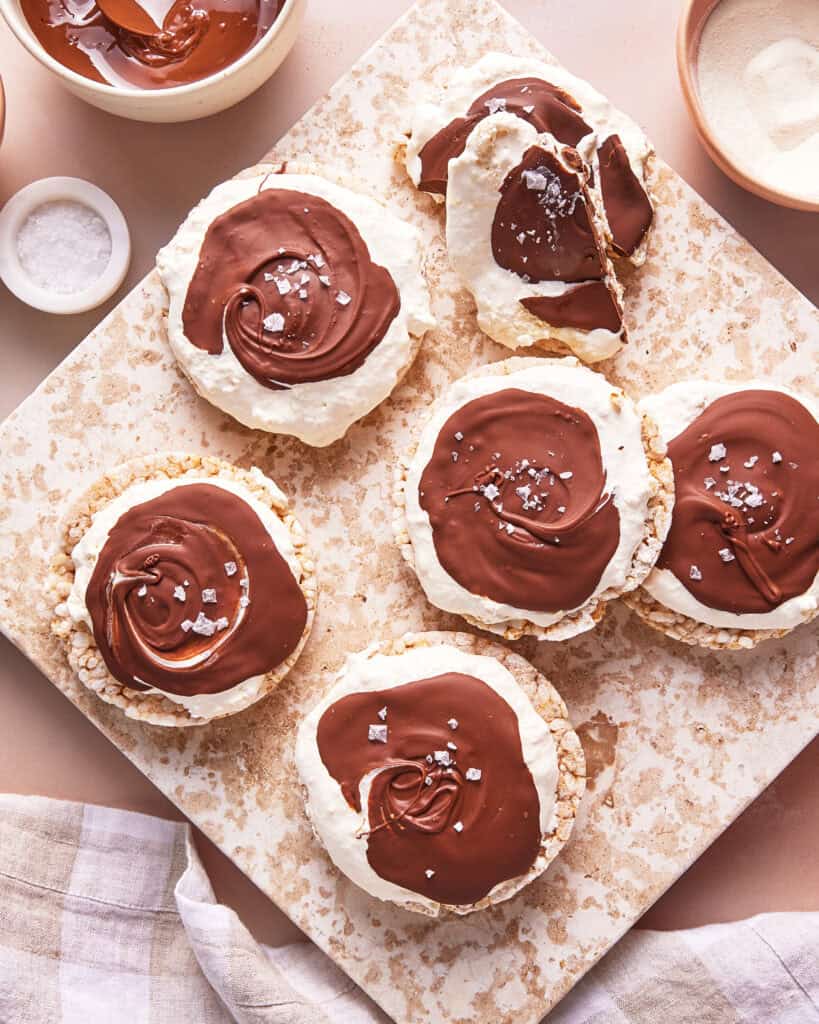 Round rice cakes topped with fluffy white cream and swirled melted chocolate, sprinkled with sea salt flakes, are arranged on a speckled cutting board. A napkin and bowls of ingredients are nearby.