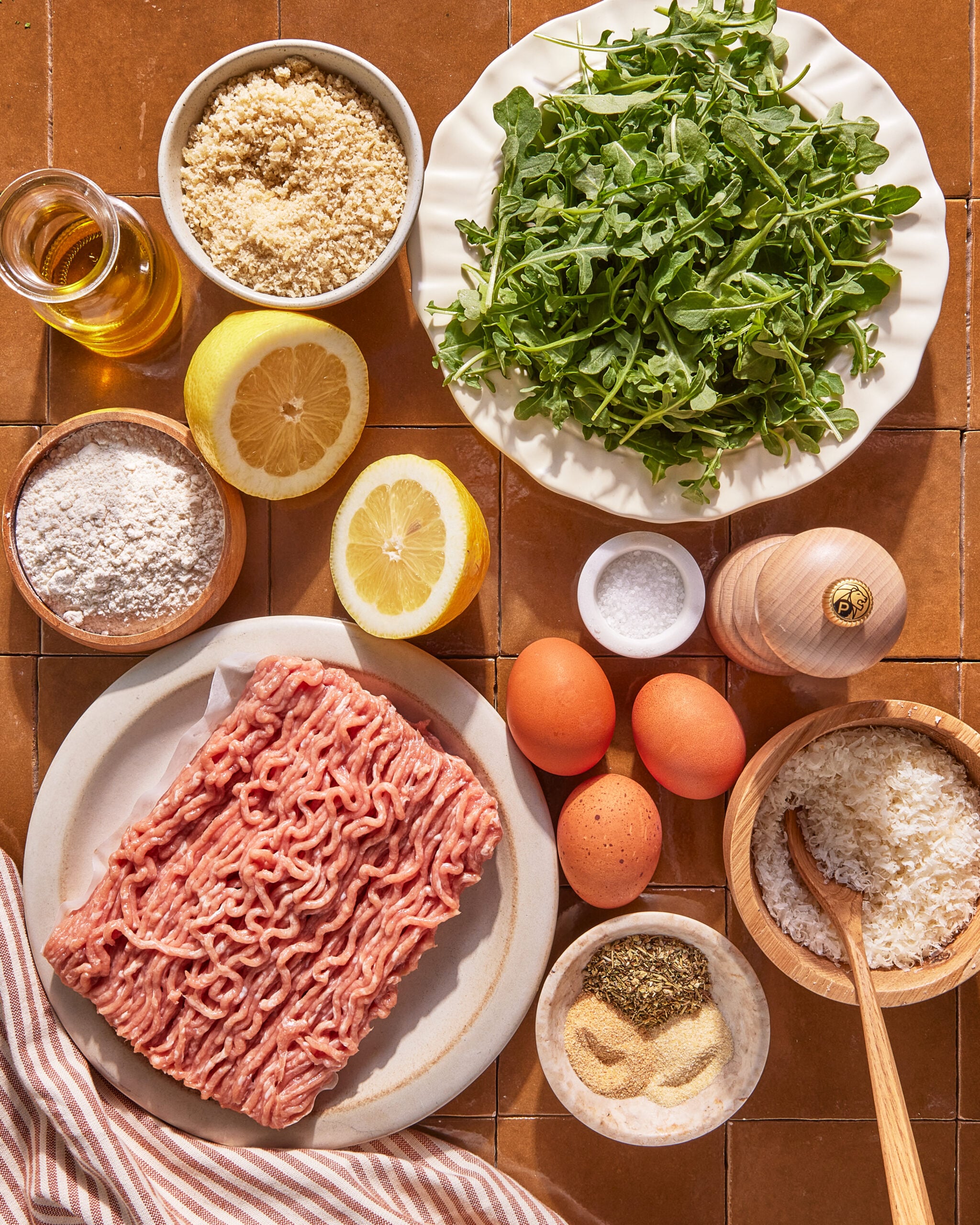 A flat lay of ingredients including ground meat, arugula, eggs, halved lemons, flour, breadcrumbs, grated cheese, olive oil, salt, pepper, and spices on a tiled surface.