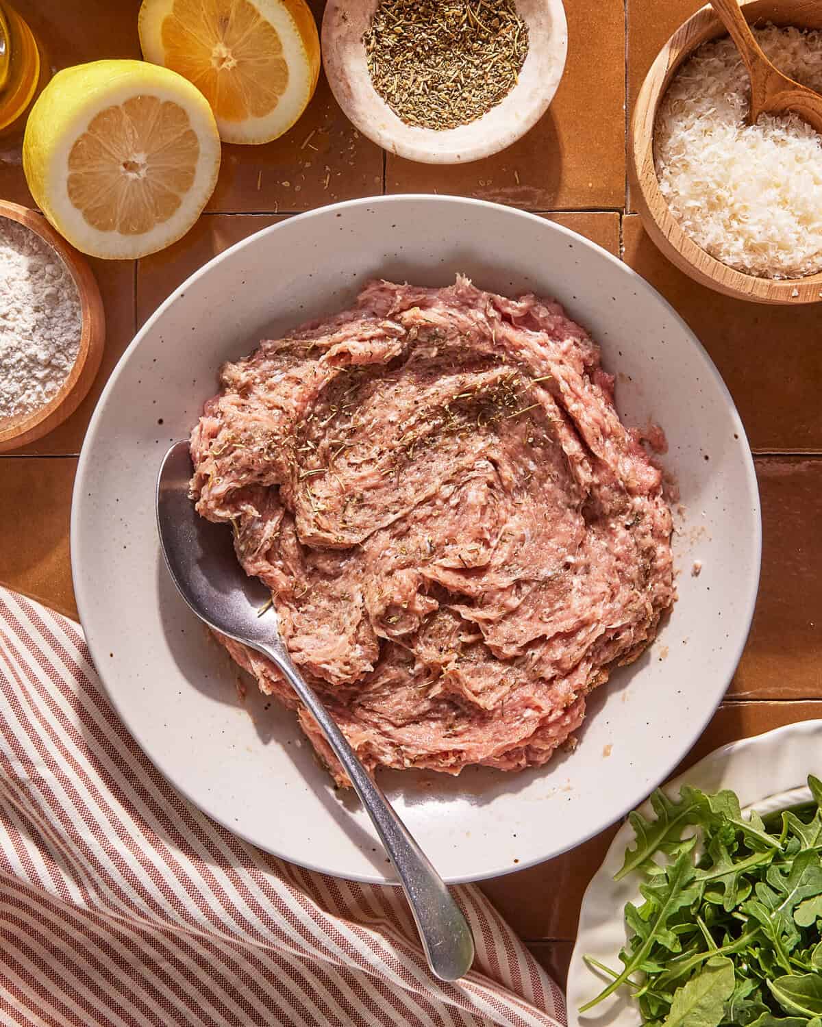 A bowl of seasoned ground meat with a spoon, surrounded by lemon halves, a bowl of herbs, grated cheese, flour, and a plate of fresh arugula on a tiled surface with a striped napkin.