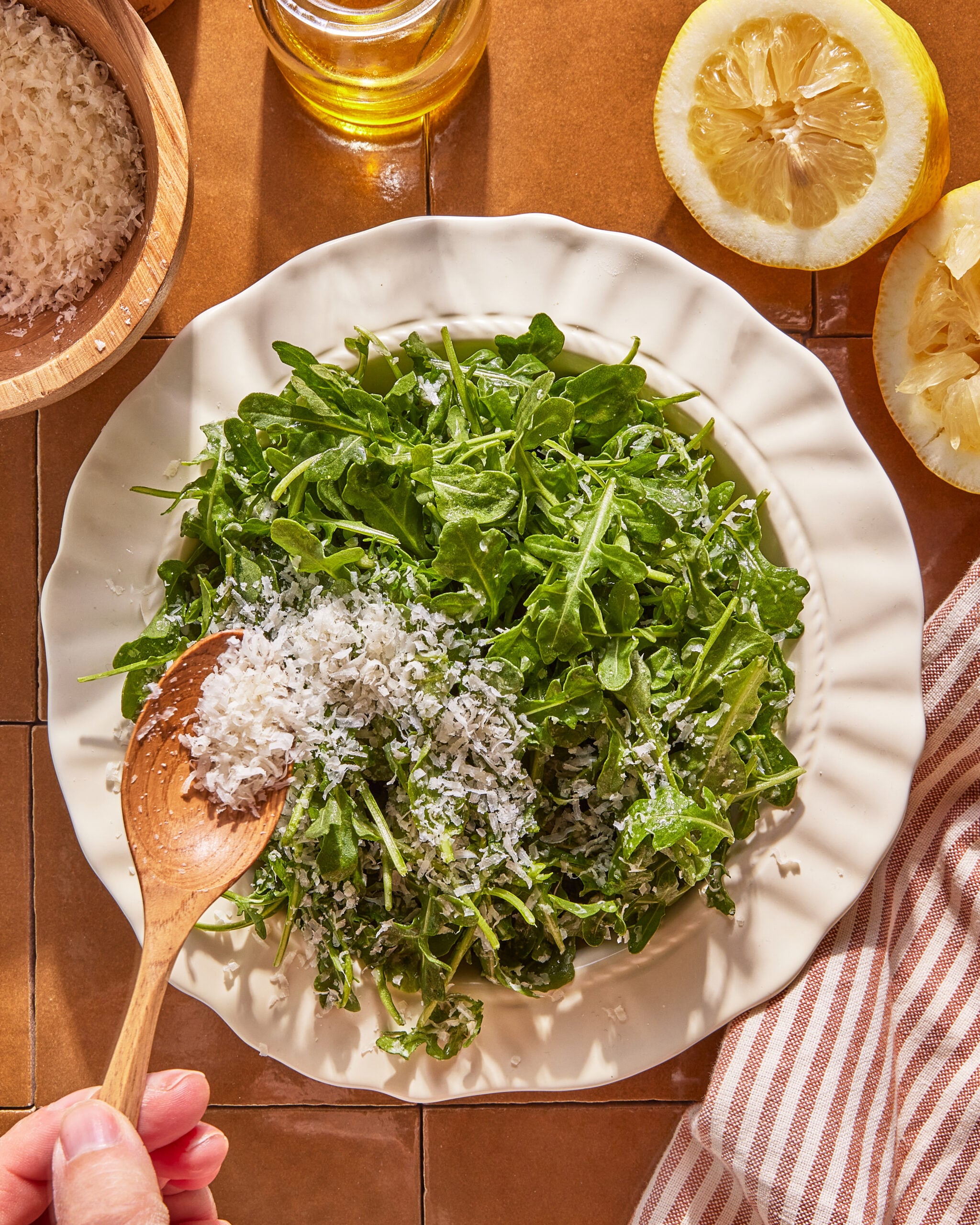 A hand sprinkles grated cheese onto a plate of fresh arugula. A halved lemon, a bowl of grated cheese, a glass of oil, and a striped towel are nearby on a brown tiled surface.
