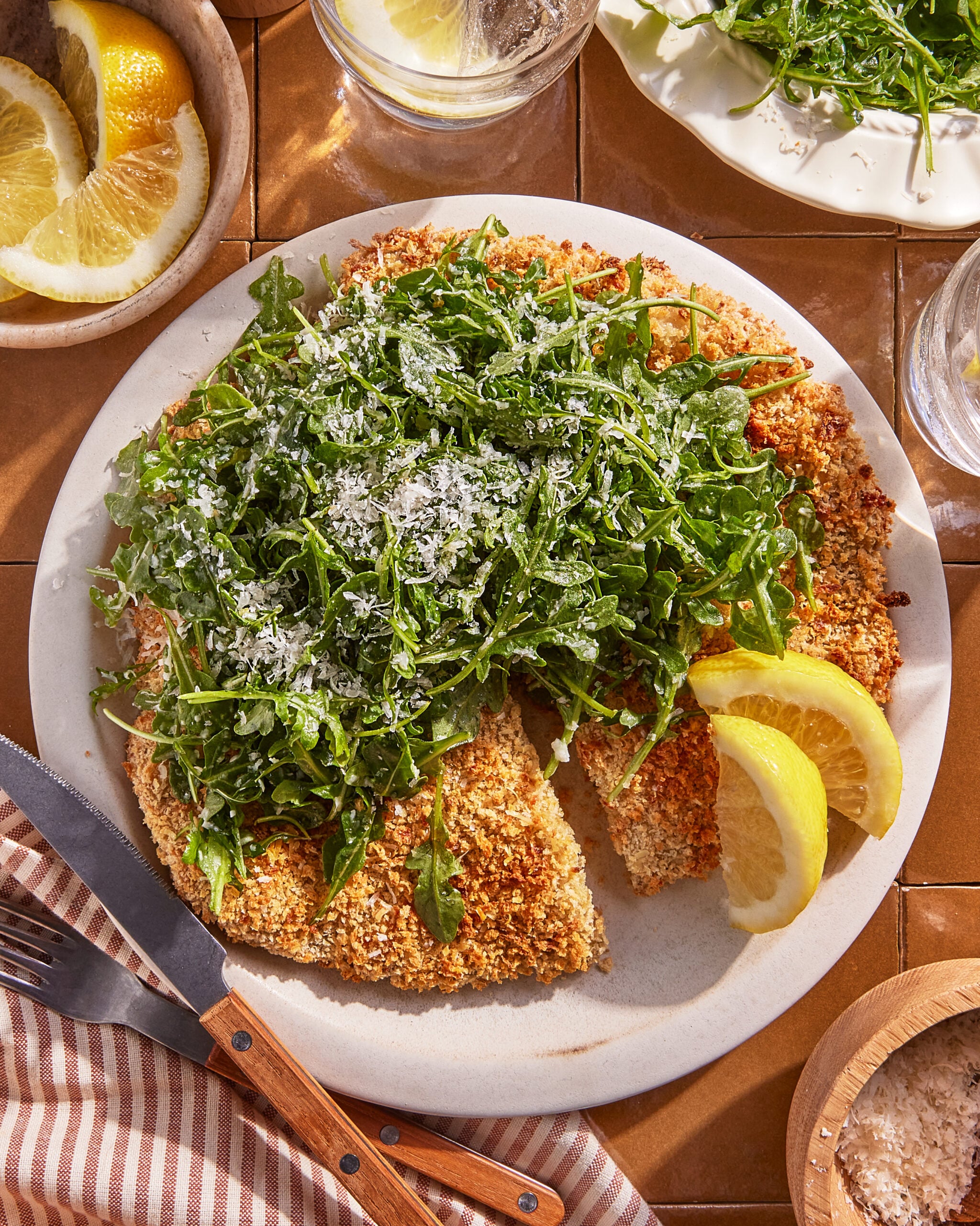 A plate of breaded chicken cutlets topped with arugula and grated cheese, garnished with lemon wedges. A fork and knife rest on the plate, and there are glasses, more arugula, and a small bowl of salt nearby.