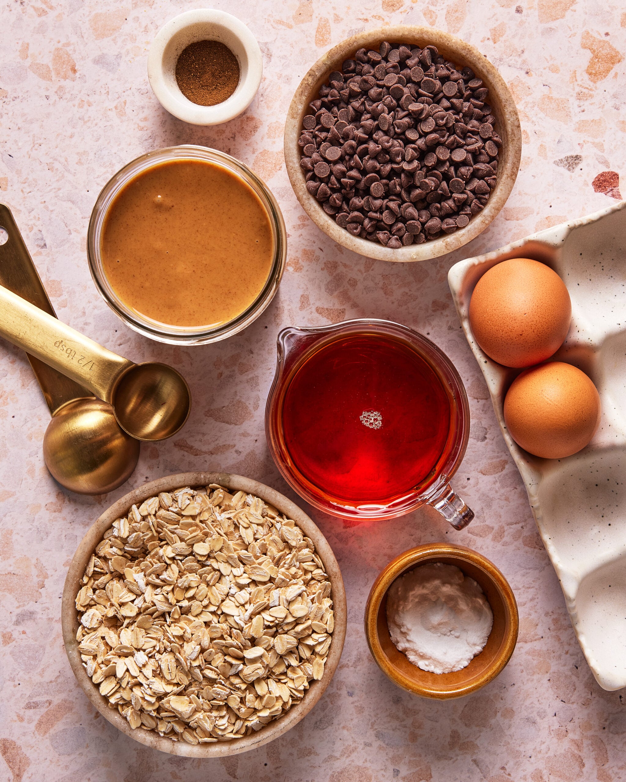 A flat lay of baking ingredients on a speckled surface, including oats, peanut butter, chocolate chips, eggs, maple syrup, baking powder, salt, and measuring spoons.
