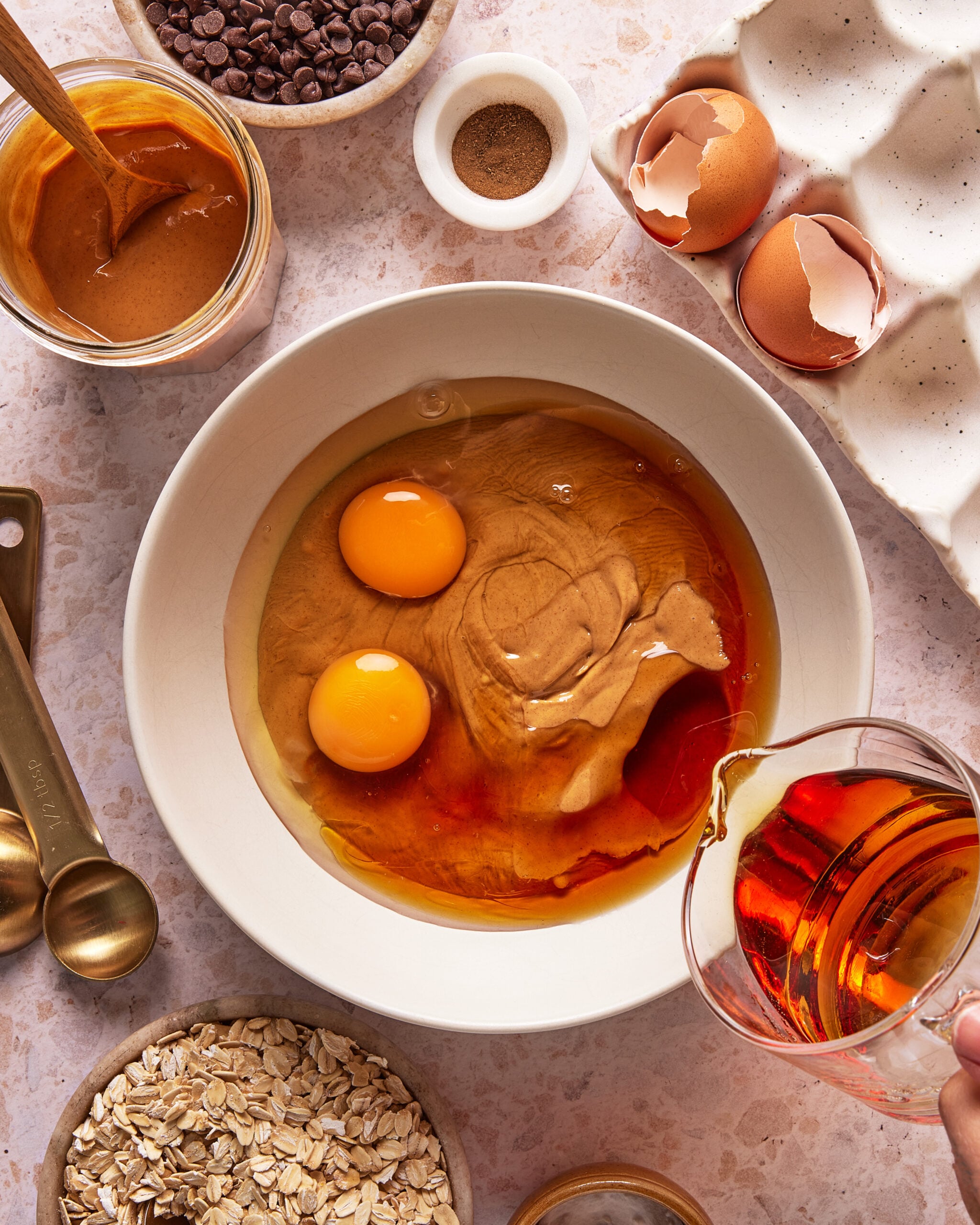 A bowl with two eggs and peanut butter is on a countertop surrounded by oats, measuring spoons, chocolate chips, and eggshells as someone pours liquid, possibly syrup or vanilla, into the bowl.