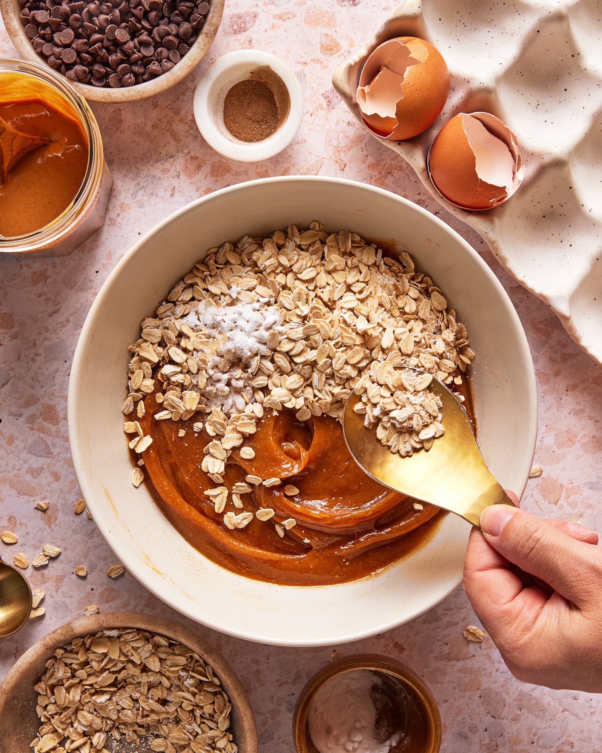 A hand holds a gold spoon above a bowl with oats, peanut butter, baking soda, and a brown mixture. Surrounding items include eggshells, chocolate chips, a jar of peanut butter, and measuring spoons on a counter.