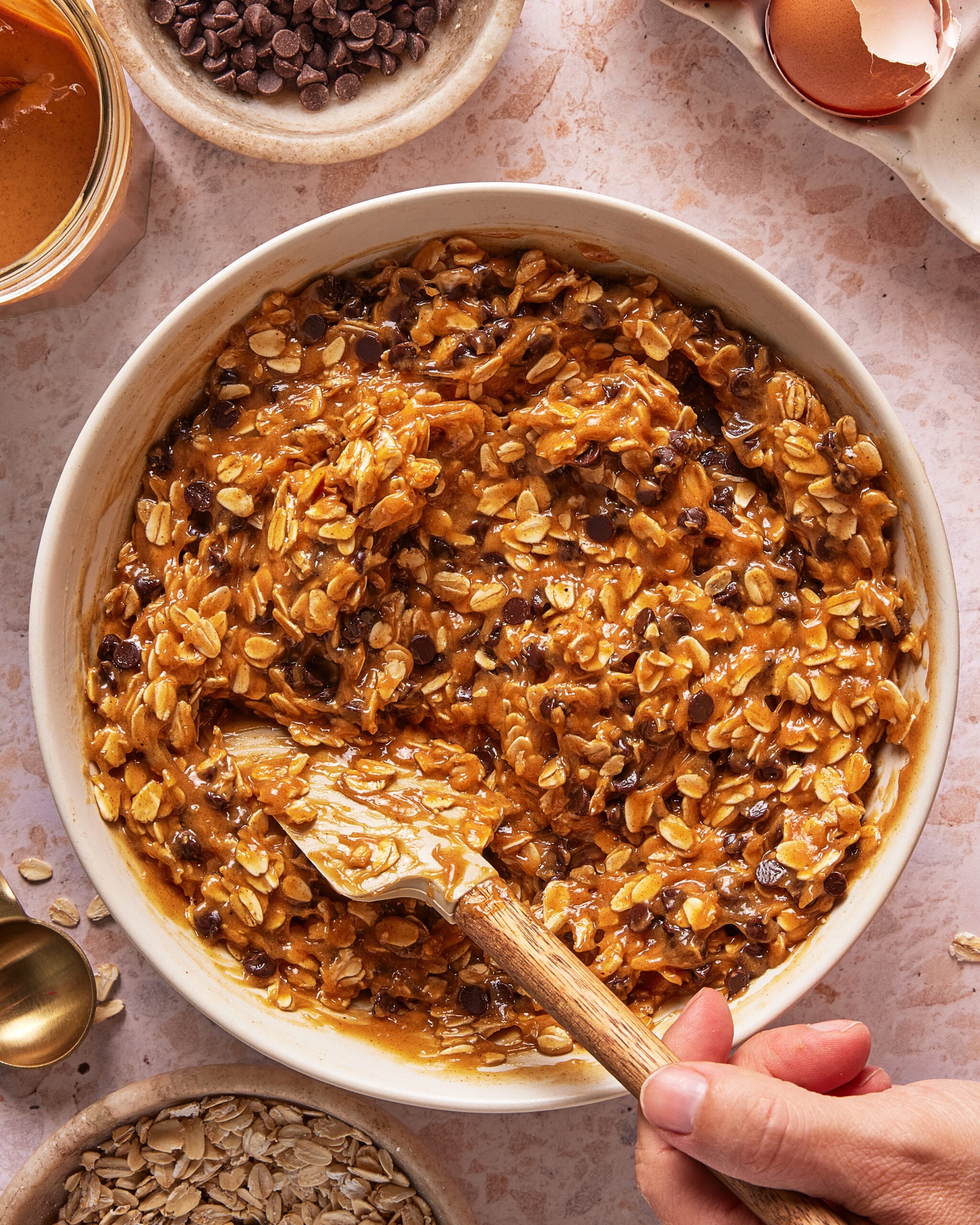 A hand stirs a mixture of oats, chocolate chips, and peanut butter in a white bowl. Ingredients like oats, chocolate chips, and a cracked egg shell are arranged around the bowl on a light countertop.