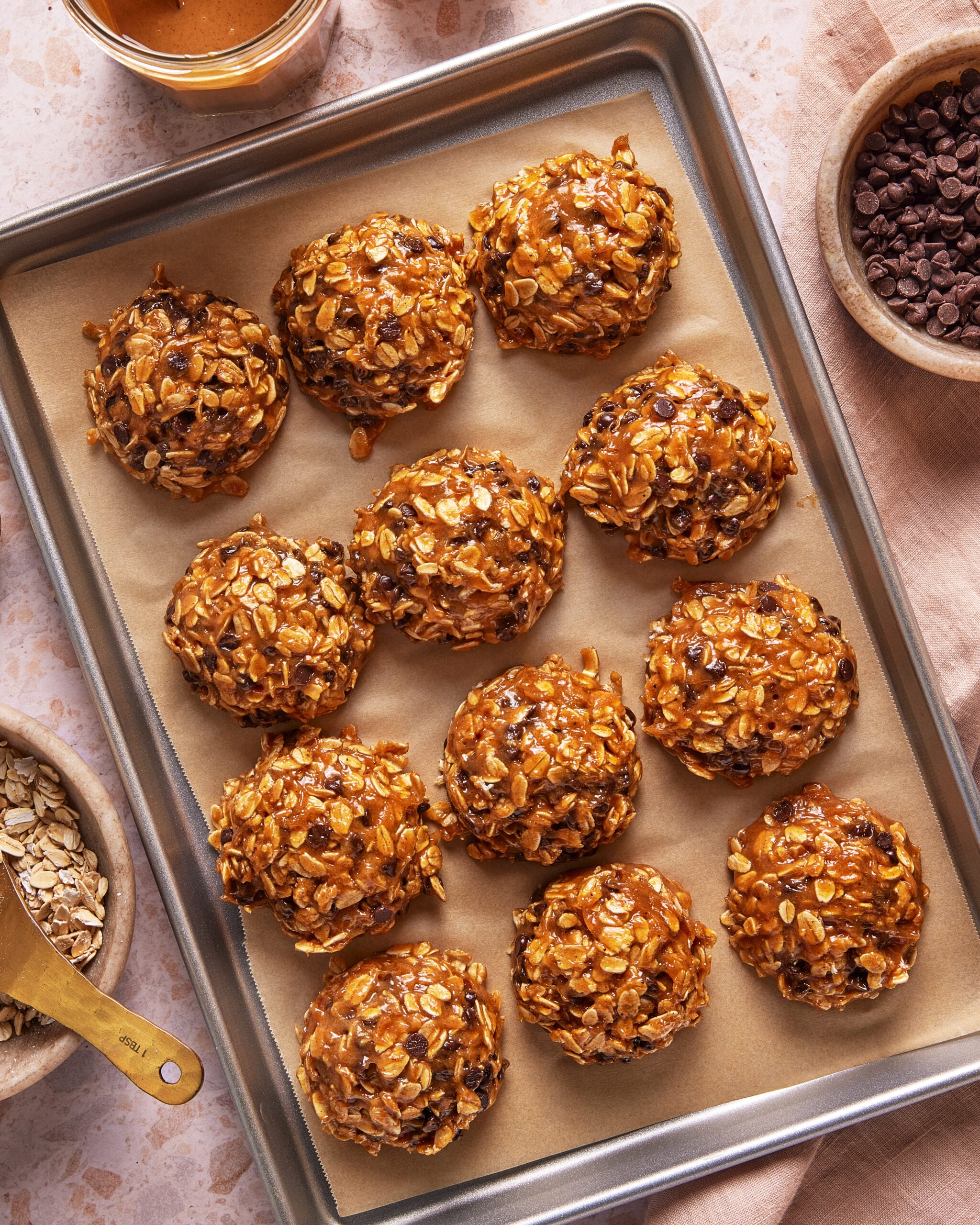 A baking sheet lined with parchment paper holds twelve round, no-bake oatmeal cookies with chocolate chips. Surrounding the tray are bowls of oats, chocolate chips, and a jar of nut butter on a light surface.