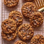 A batch of oatmeal chocolate chip cookies topped with flaky sea salt cooling on a wire rack, with a gold spatula nearby.