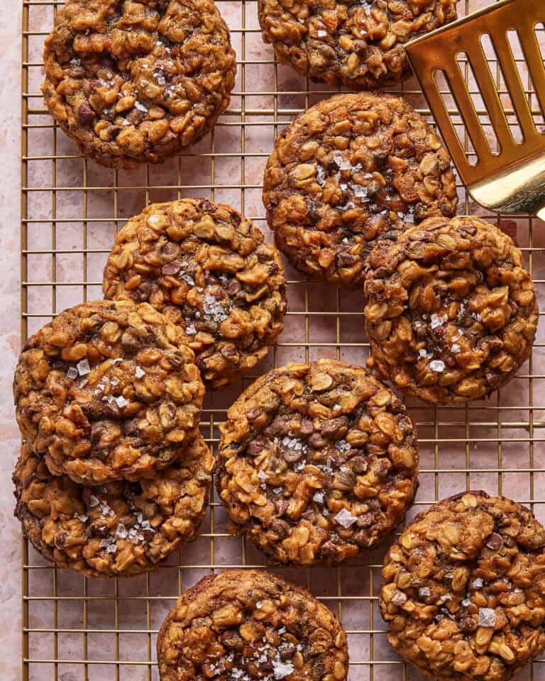 A batch of oatmeal chocolate chip cookies topped with flaky sea salt cooling on a wire rack, with a gold spatula nearby.