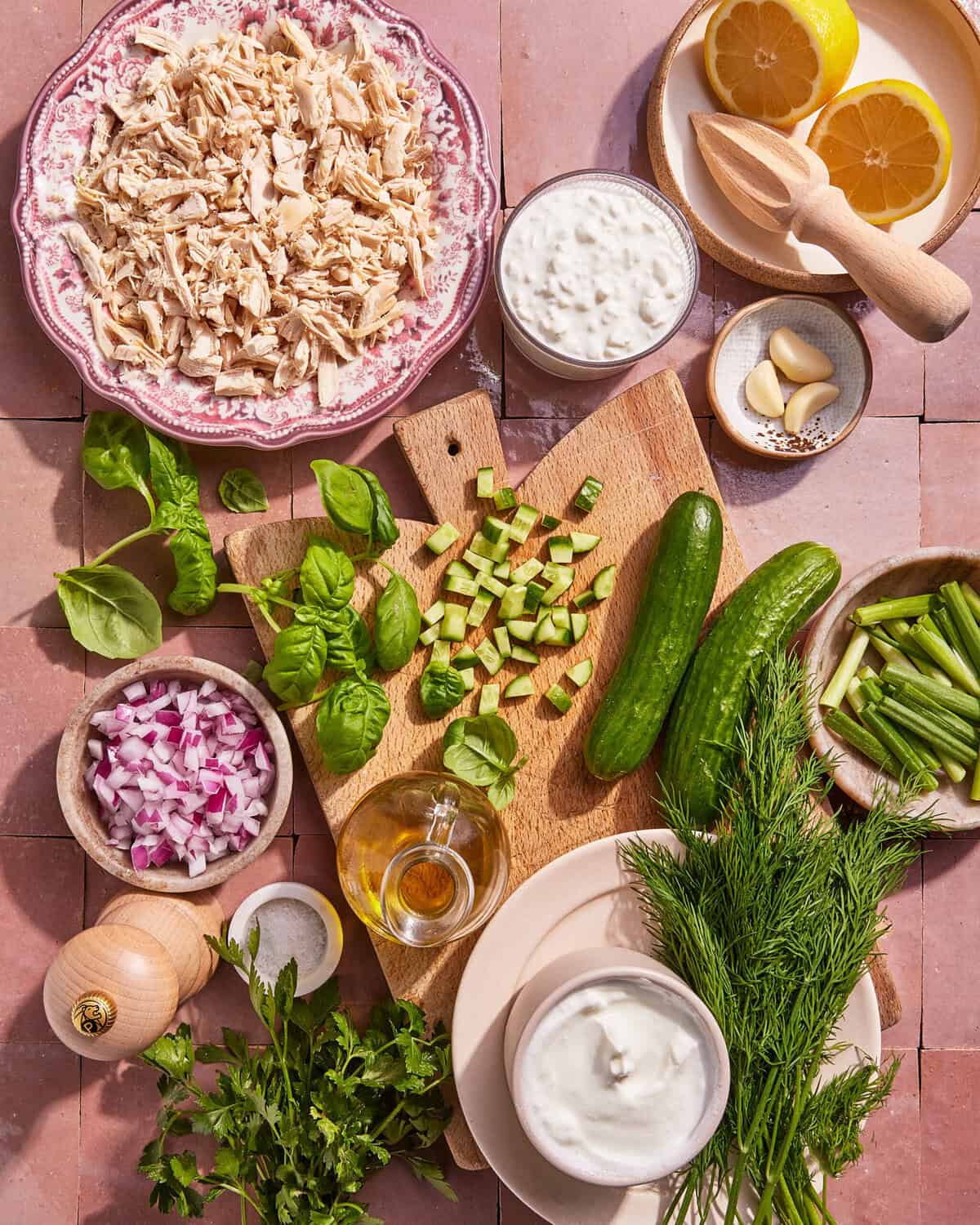 A flat lay of ingredients on a pink tiled surface, including shredded chicken, chopped cucumber, red onion, green onions, fresh herbs, cucumber, lemon halves, olive oil, garlic cloves, and bowls of yogurt and cottage cheese.
