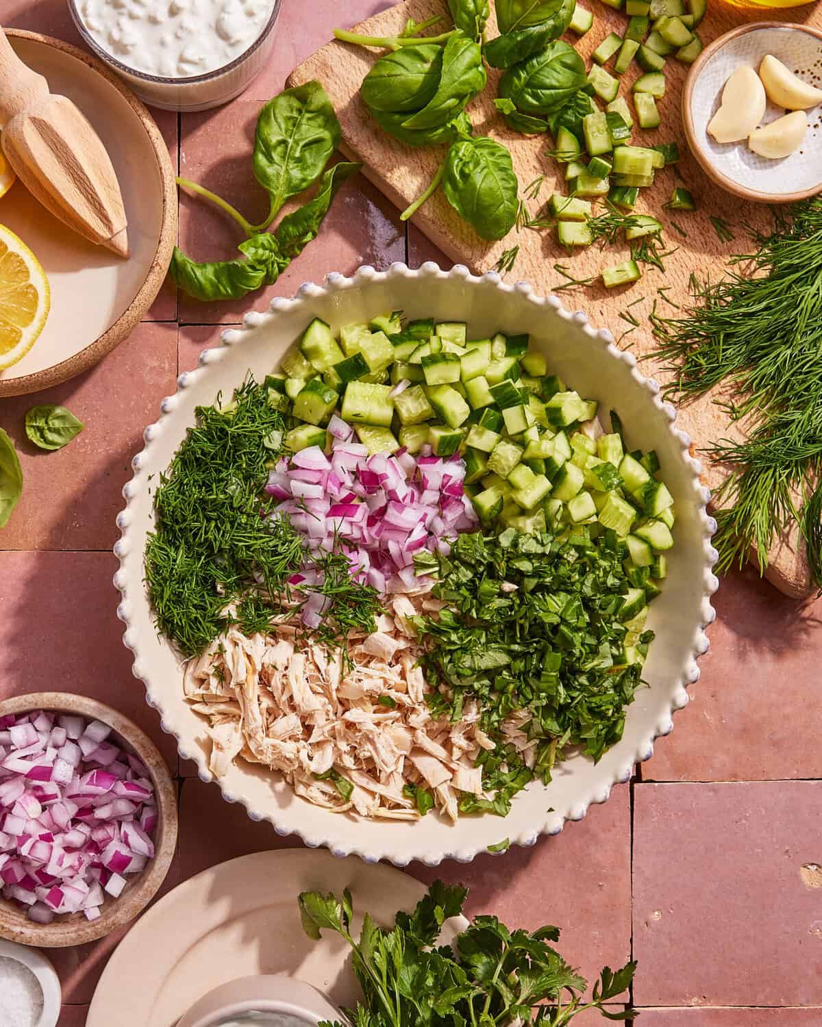 A bowl containing chopped cucumber, red onion, dill, herbs, and shredded chicken sits on a pink tile surface surrounded by fresh greens, garlic, lemon, and small bowls of ingredients.