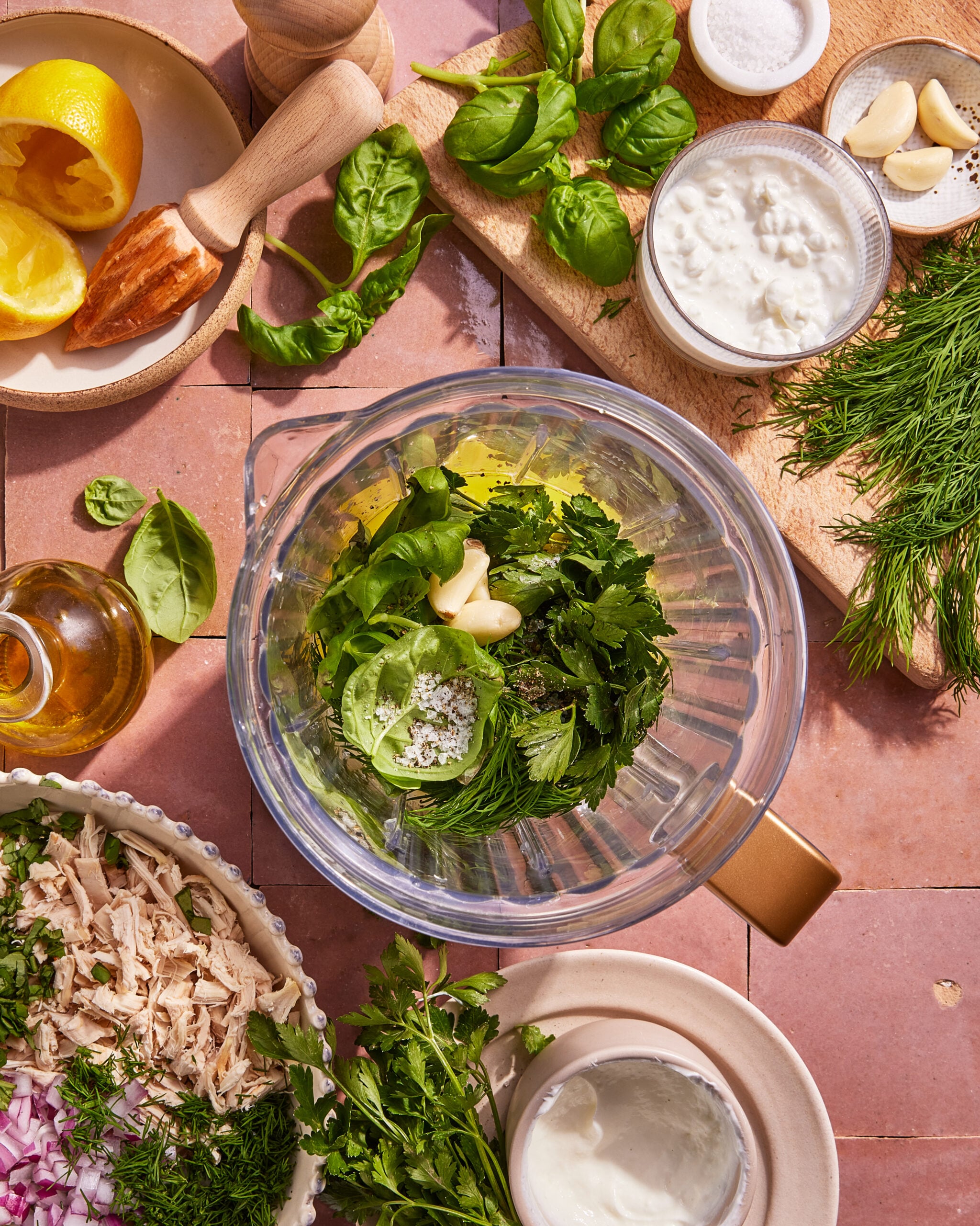 A blender filled with fresh herbs, garlic, and salt sits on a tiled surface, surrounded by bowls of shredded chicken, yogurt, herbs, lemon, olive oil, and a wooden citrus reamer.