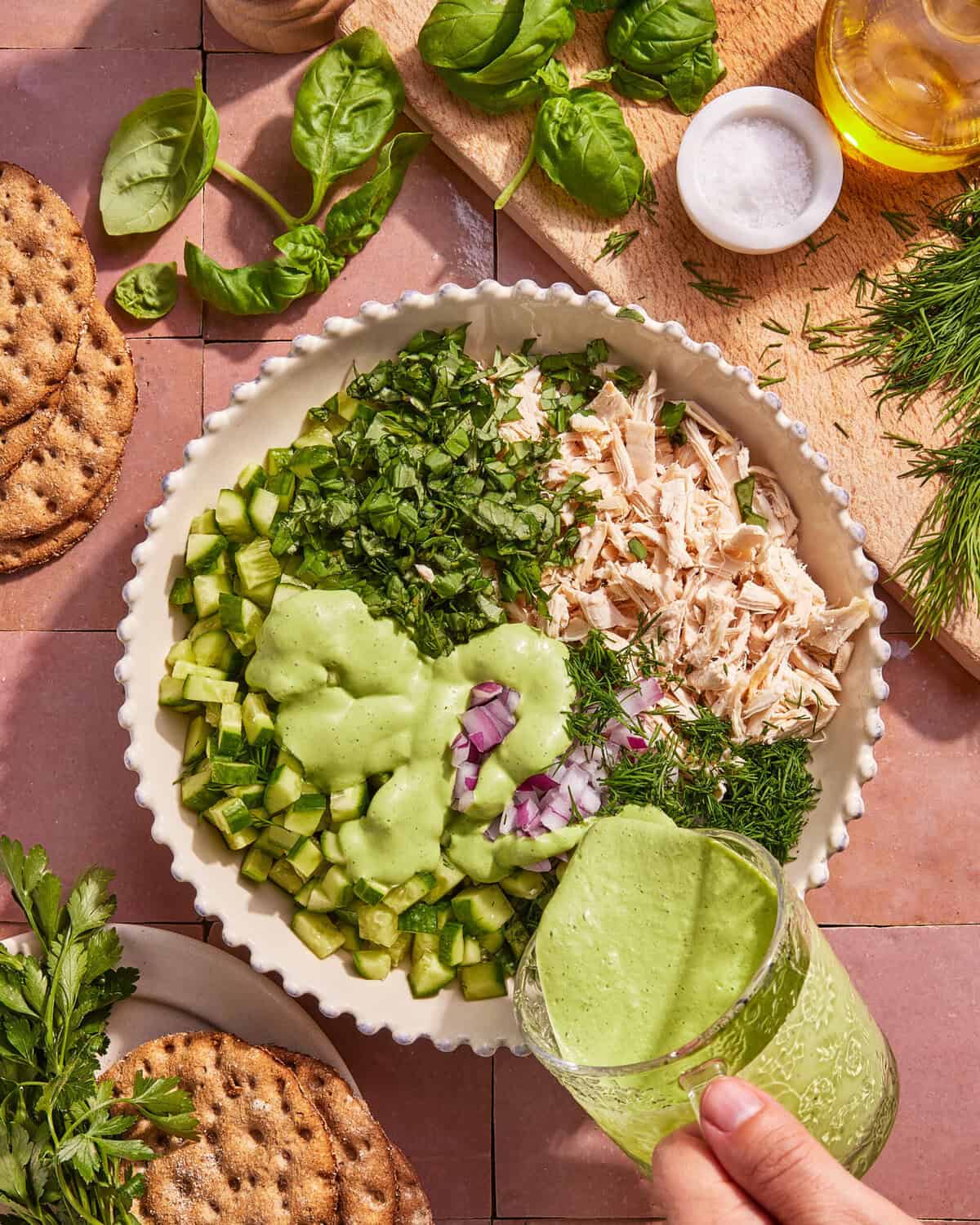 A hand pours green dressing into a bowl filled with diced cucumber, shredded chicken, chopped herbs, and red onion. Crackers, herbs, olive oil, and salt are arranged around the bowl on a pink tile surface.