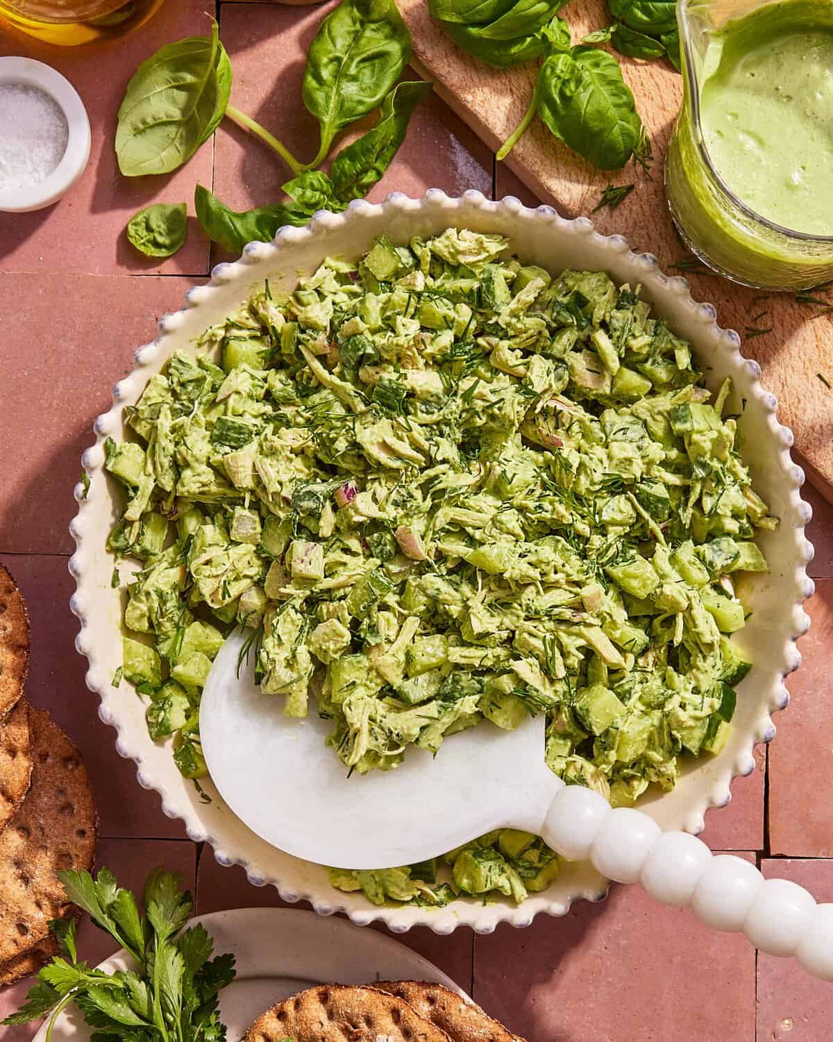 A white bowl filled with a chopped green salad mixture, including herbs and vegetables, sits on a tile surface. A white serving spoon rests in the bowl, with crackers, basil leaves, and green dressing nearby.