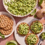 Three crispbreads topped with a green chopped salad sit on a plate, with extra crispbreads, a large bowl of salad, a jug of green dressing, fresh herbs, and a pepper grinder nearby on a tiled surface.