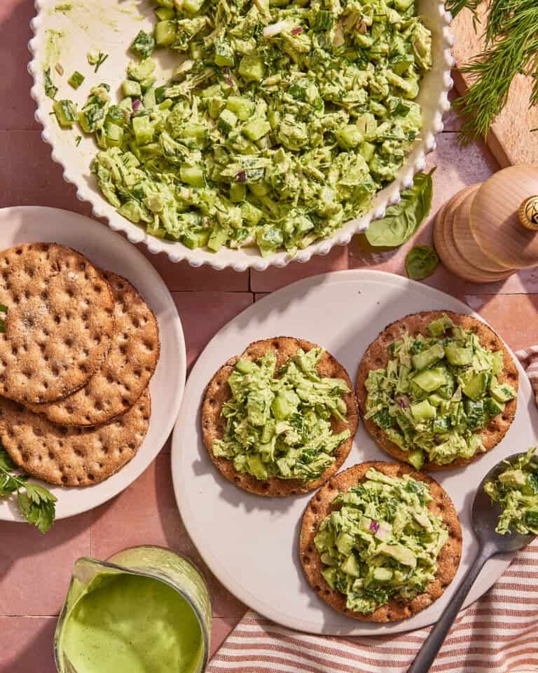Three crispbreads topped with a green chopped salad sit on a plate, with extra crispbreads, a large bowl of salad, a jug of green dressing, fresh herbs, and a pepper grinder nearby on a tiled surface.